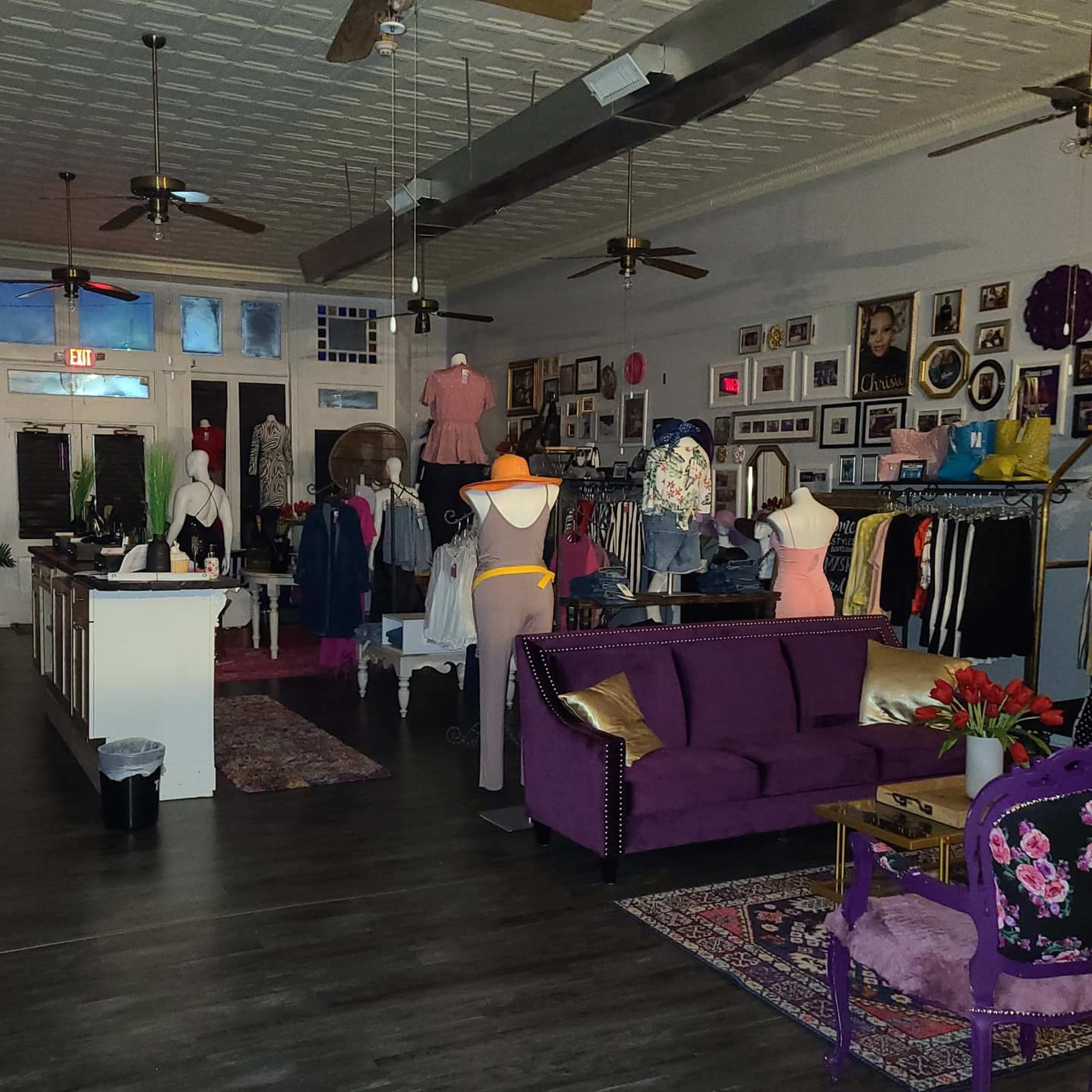 A clothing store features mannequins dressed in various outfits, a purple sofa, and framed pictures lining the walls.