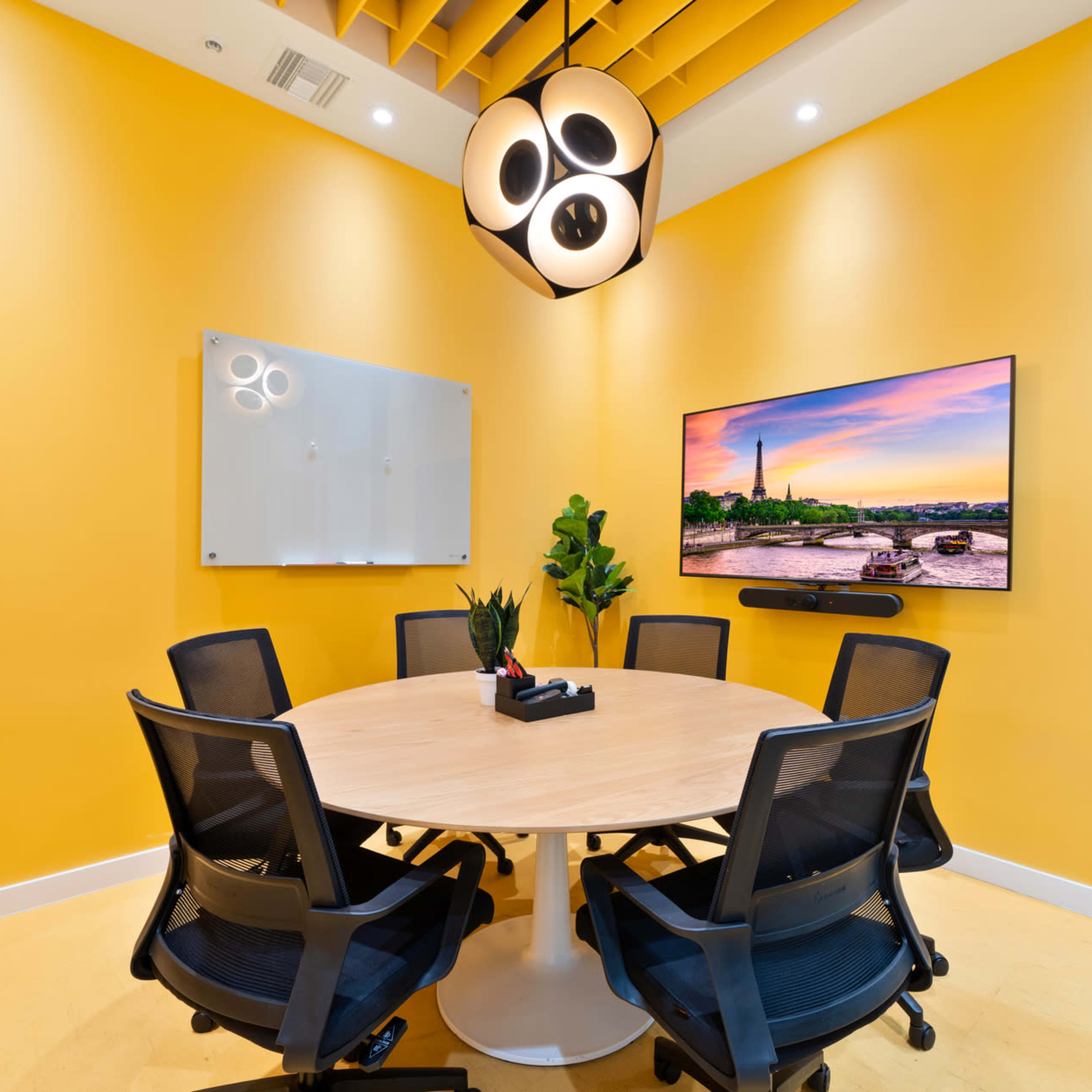 A brightly colored meeting room features a round wooden table surrounded by six black chairs, with a large screen displaying a scenic image on the wall.