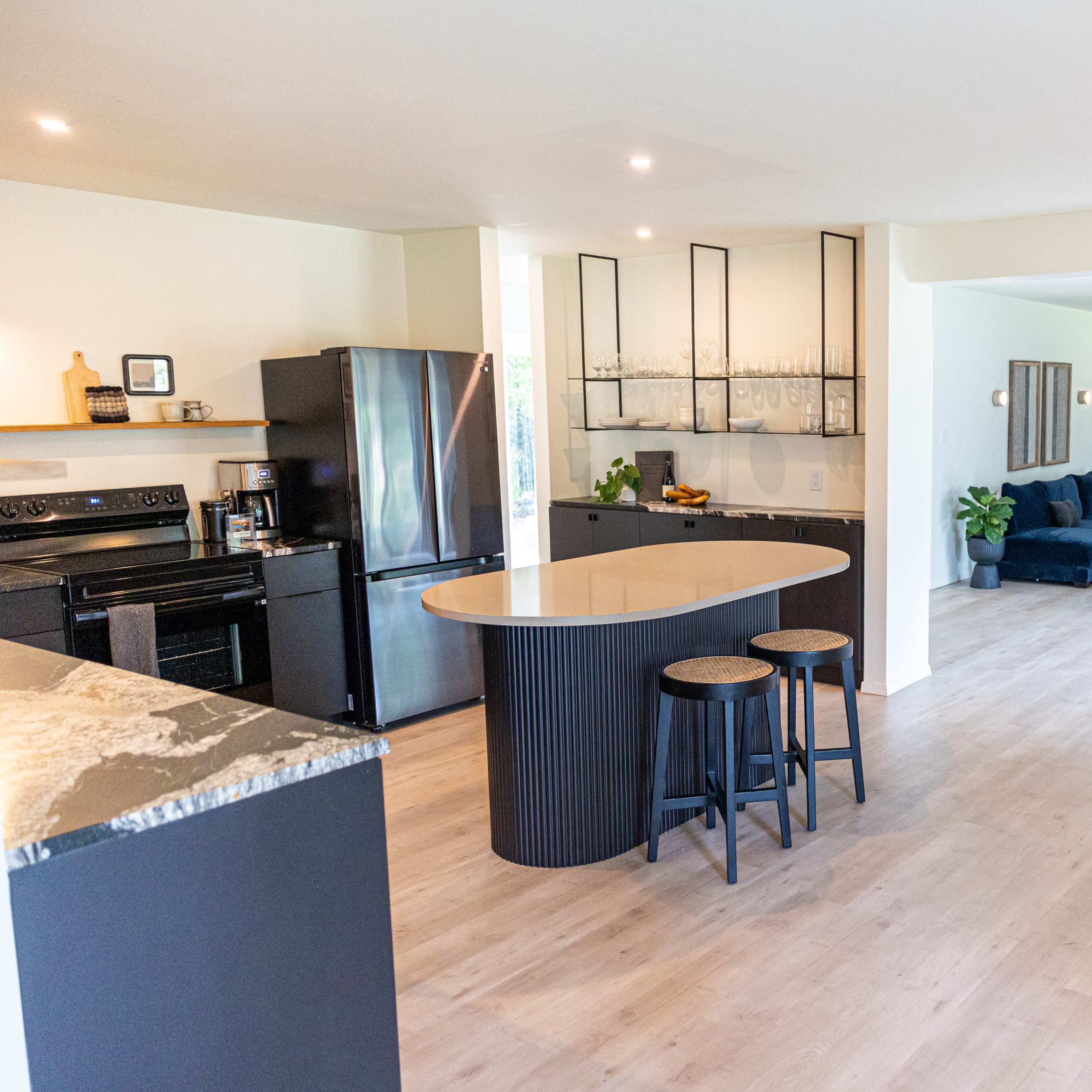 A modern, open-concept kitchen and living area featuring sleek black appliances, a central island with bar stools, and a large sectional sofa placed near a stacked log feature.