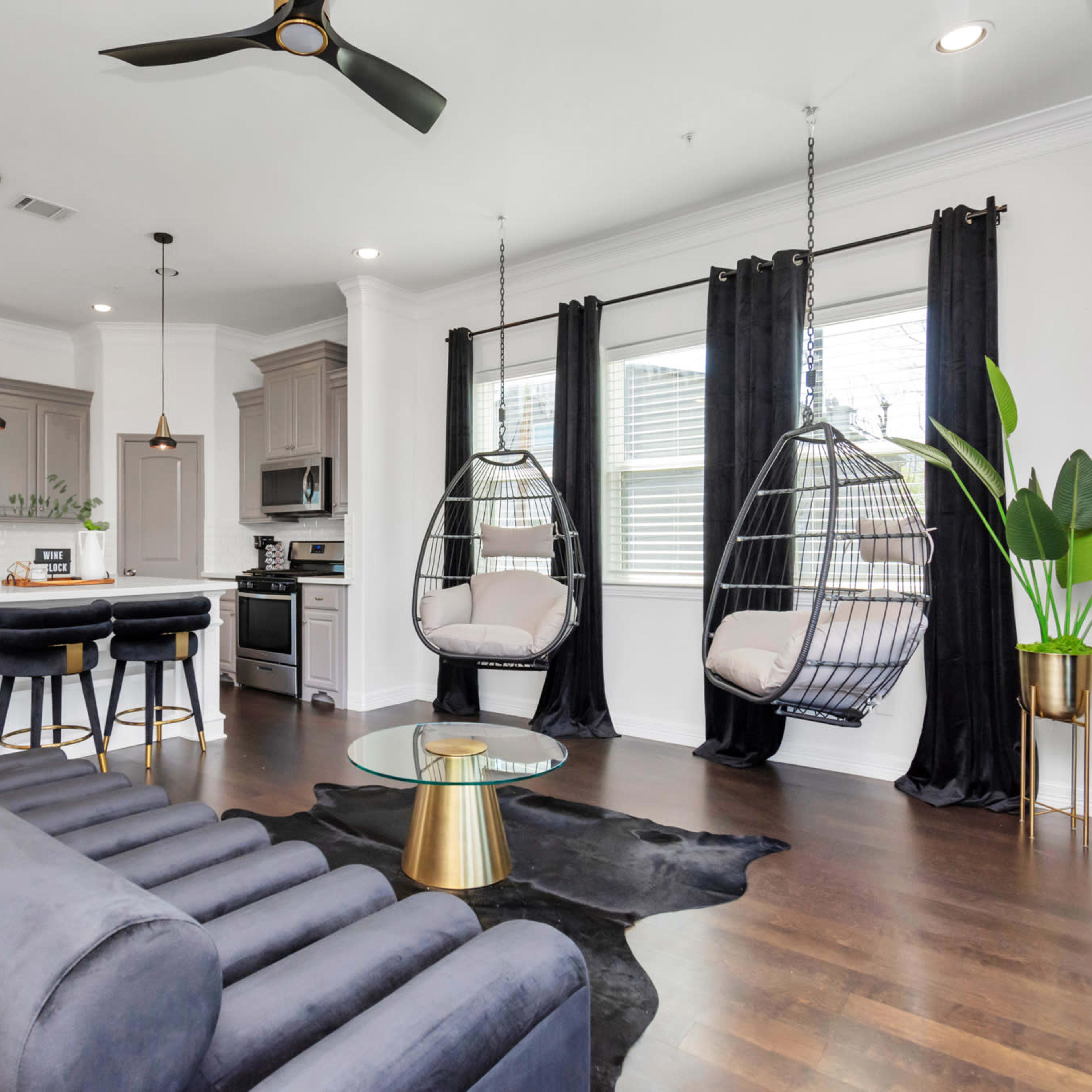 A modern living area with a gray sofa, two hanging chairs, a glass coffee table, and a kitchen in the background.