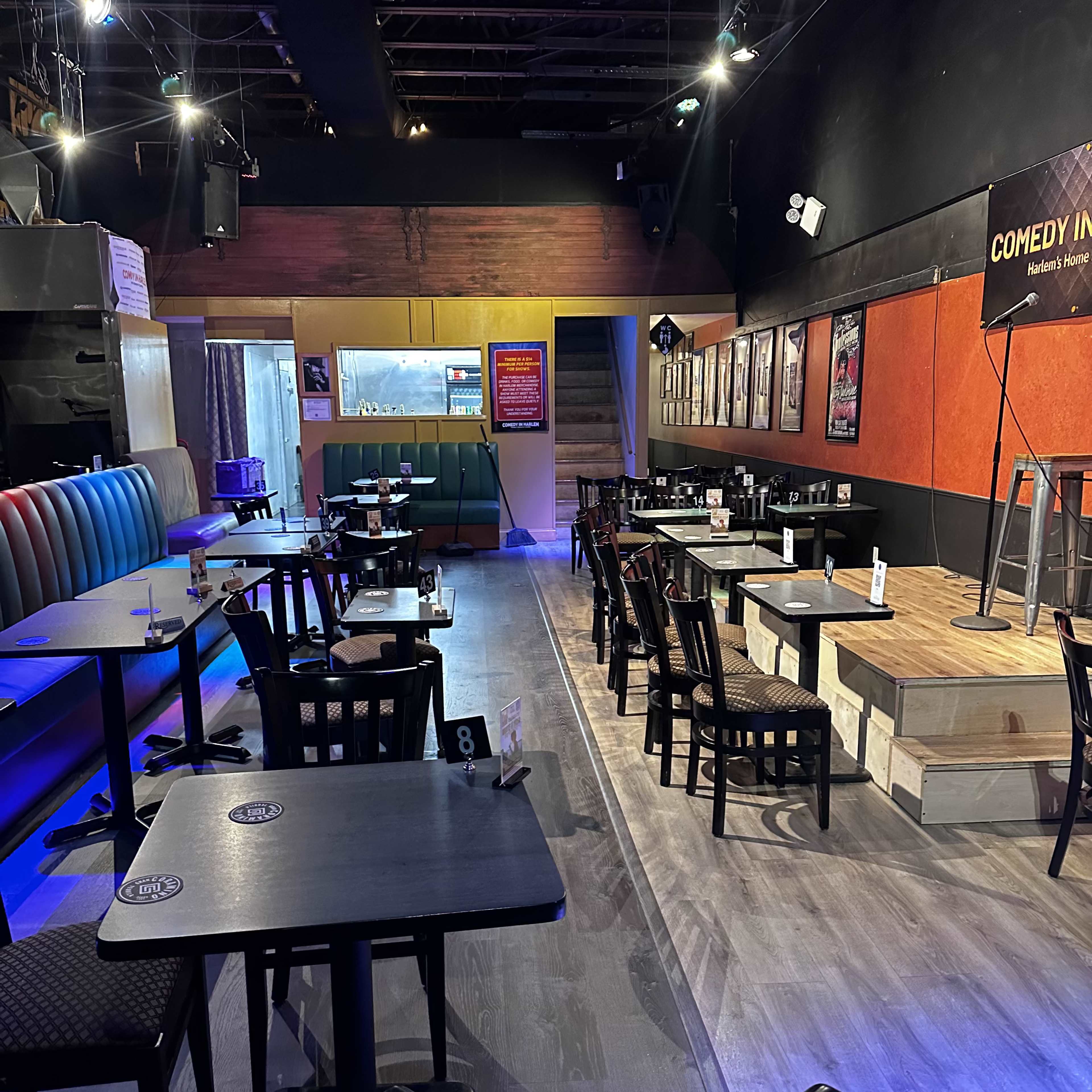 A dimly lit comedy club with empty tables and a small stage at the front.