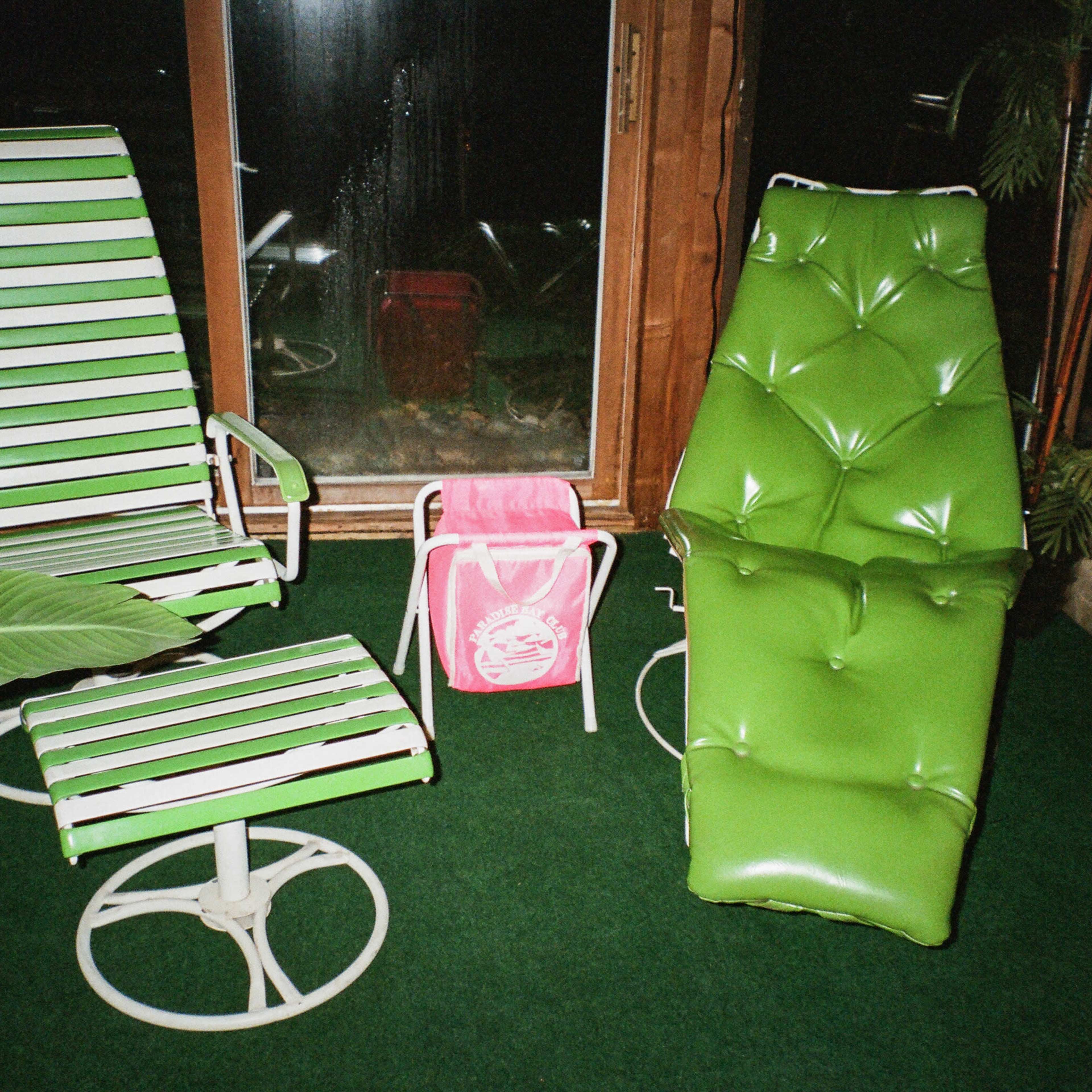 Two green lounge chairs, a small green footrest, and a pink bag, set against a backdrop of large windows and indoor plants on a carpeted floor.