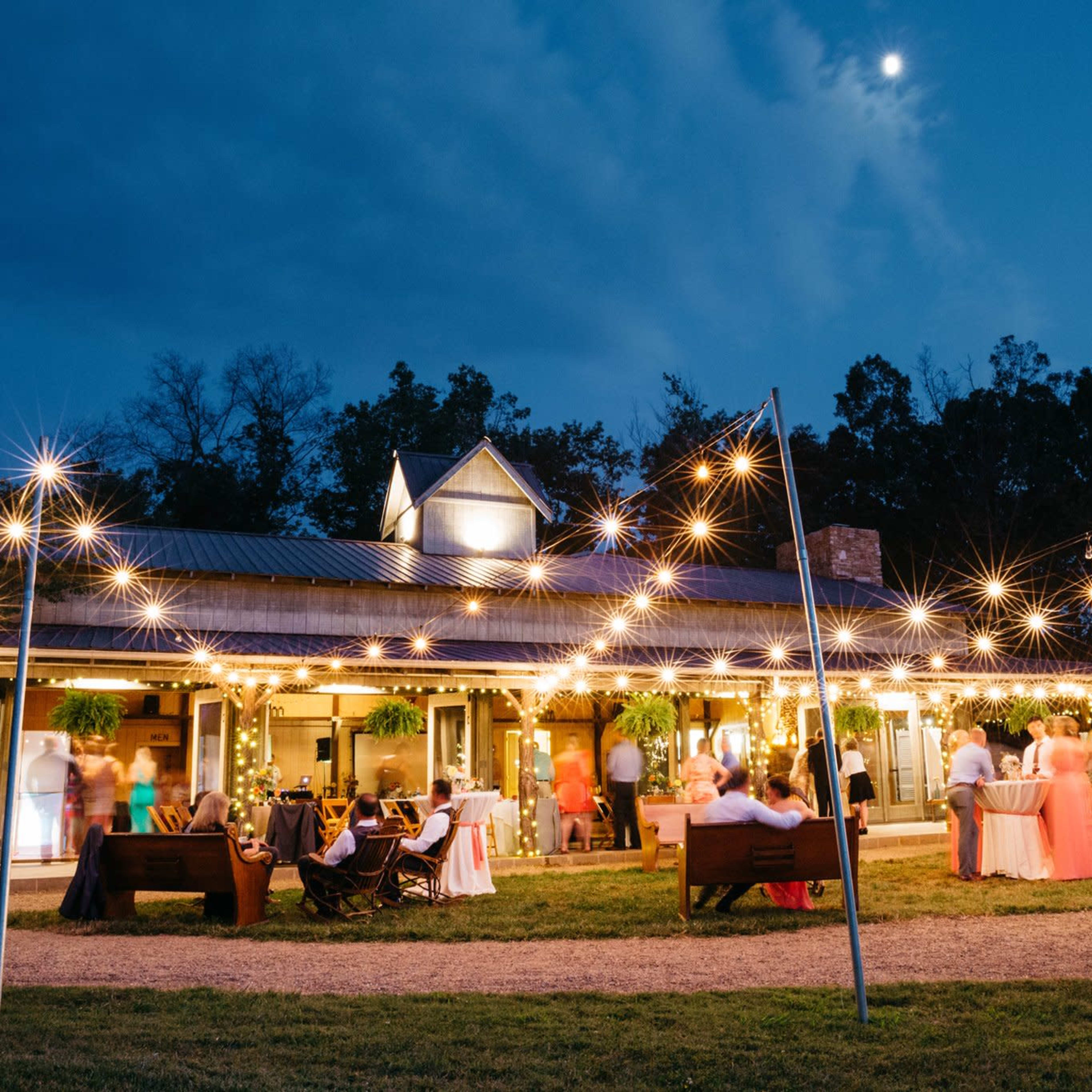 A gathering is taking place outdoors at a venue illuminated by string lights, with guests mingling near tables and a clear sky visible at dusk.