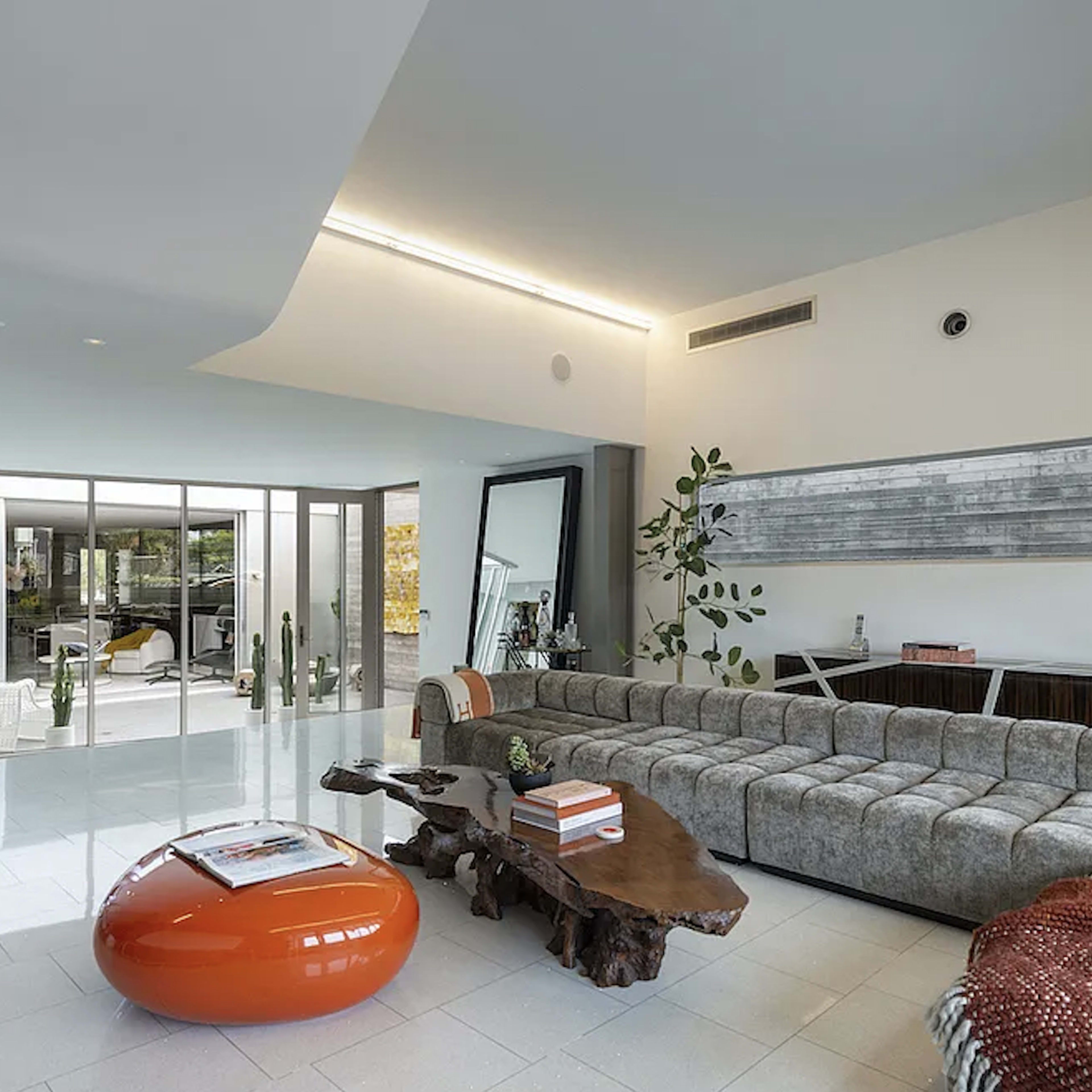 A modern living room with a large sectional sofa, a wooden coffee table, an orange round ottoman, and floor-to-ceiling windows that lead to an outdoor space.