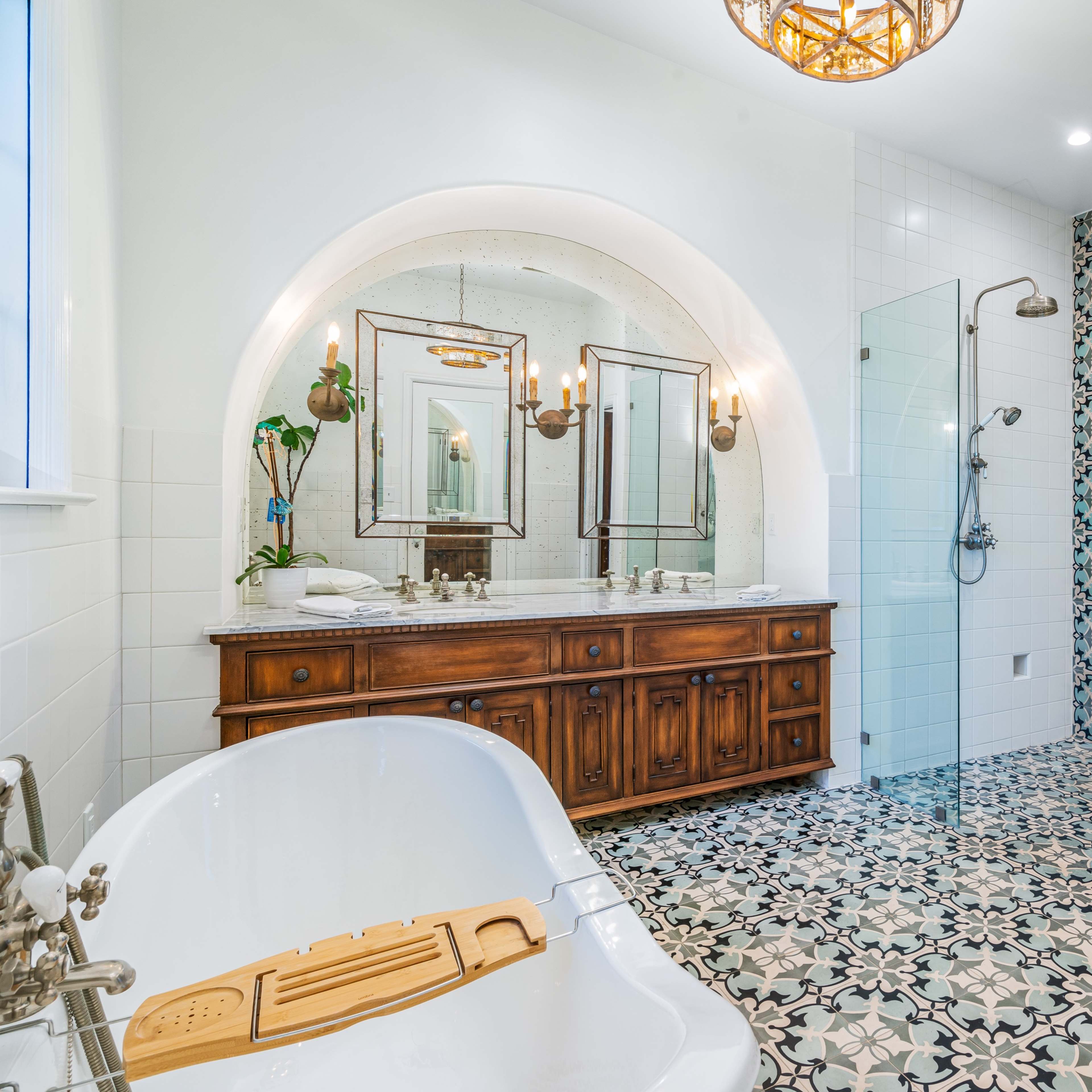 The bathroom features a freestanding tub in front of a large arched mirror, a glass shower enclosure, and intricate patterned tile flooring.