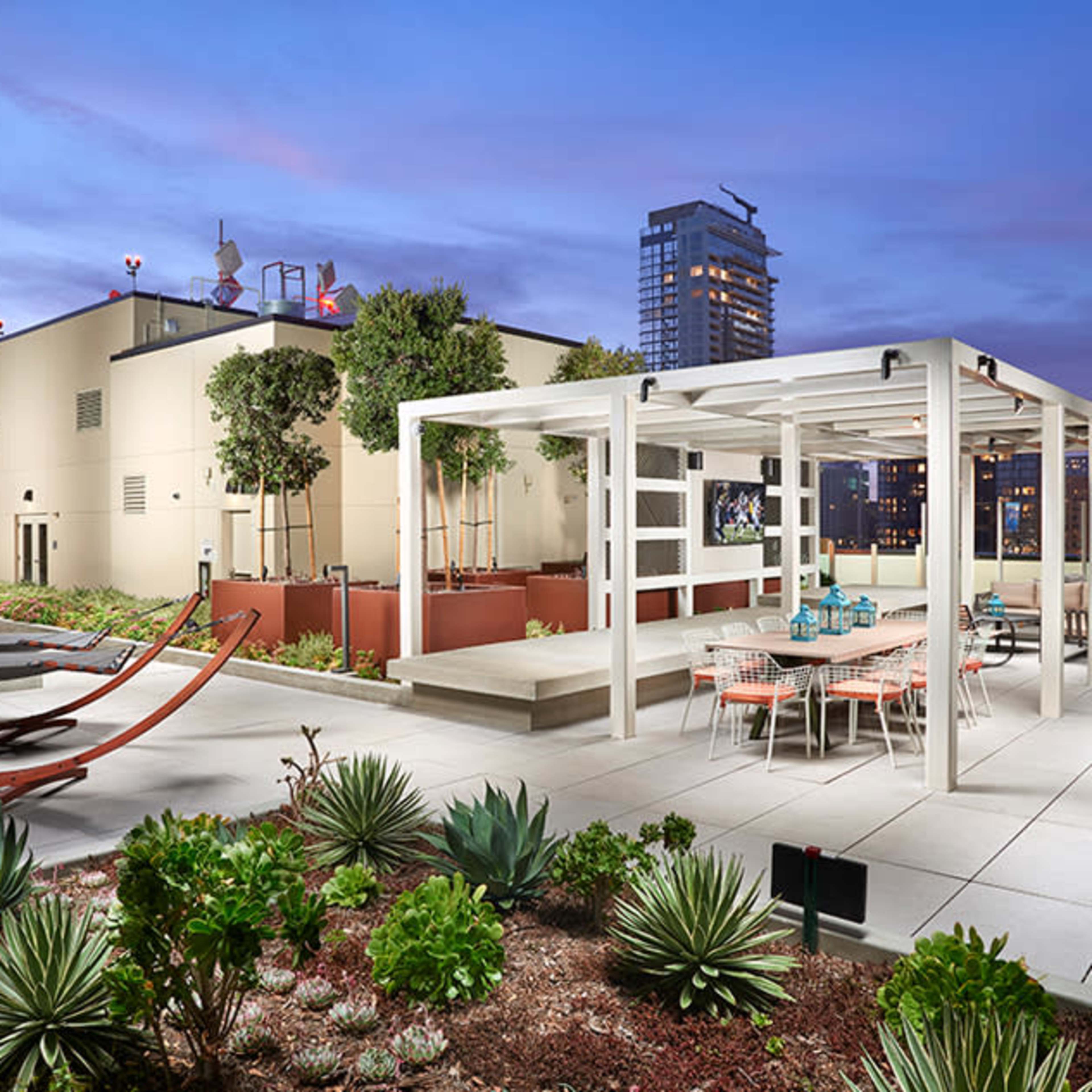 A rooftop terrace featuring a pergola, seating area with tables and chairs, and landscaped plants under a twilight sky.