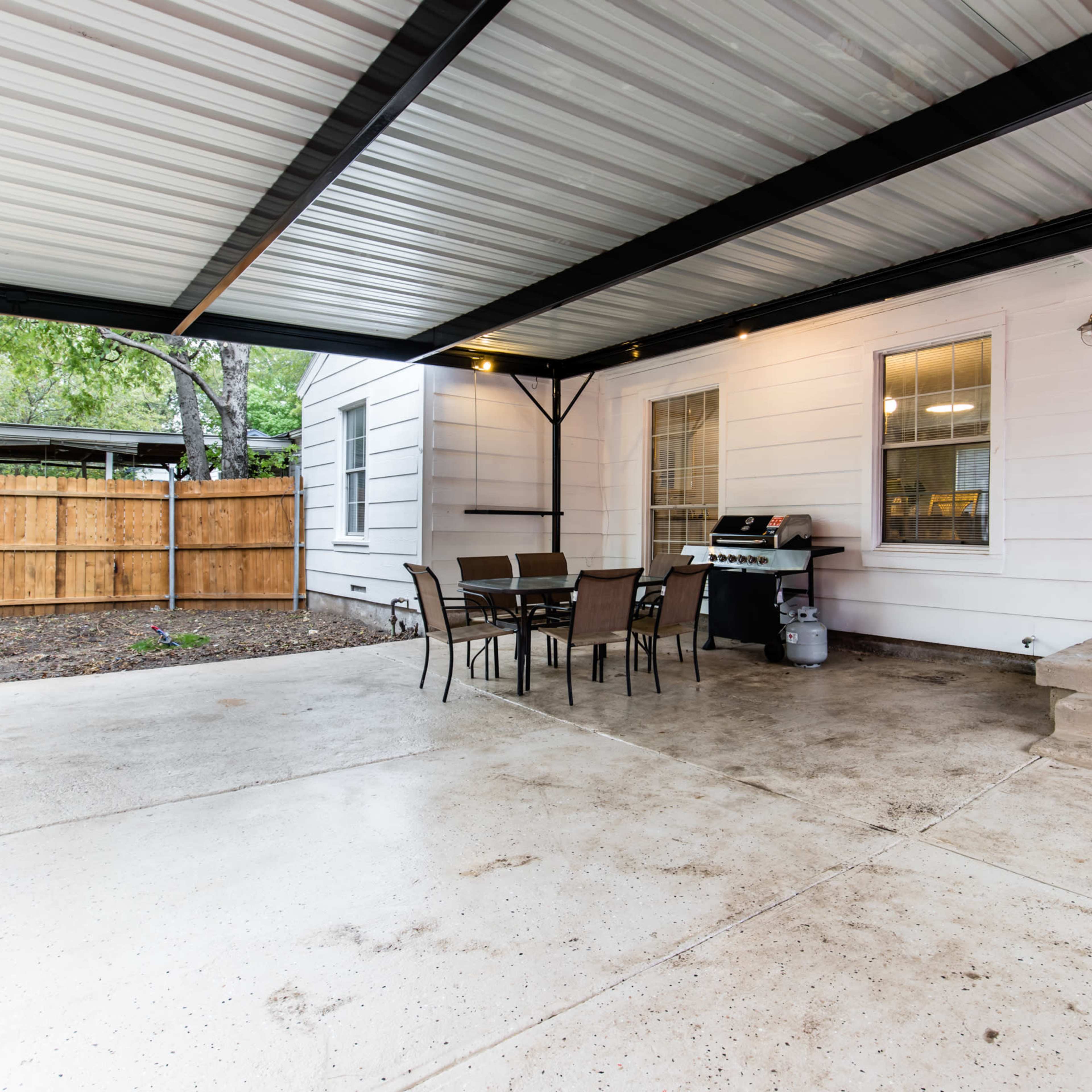 A covered patio area with a concrete floor, a table and chairs, and a grill, adjacent to a house and a wooden fence.