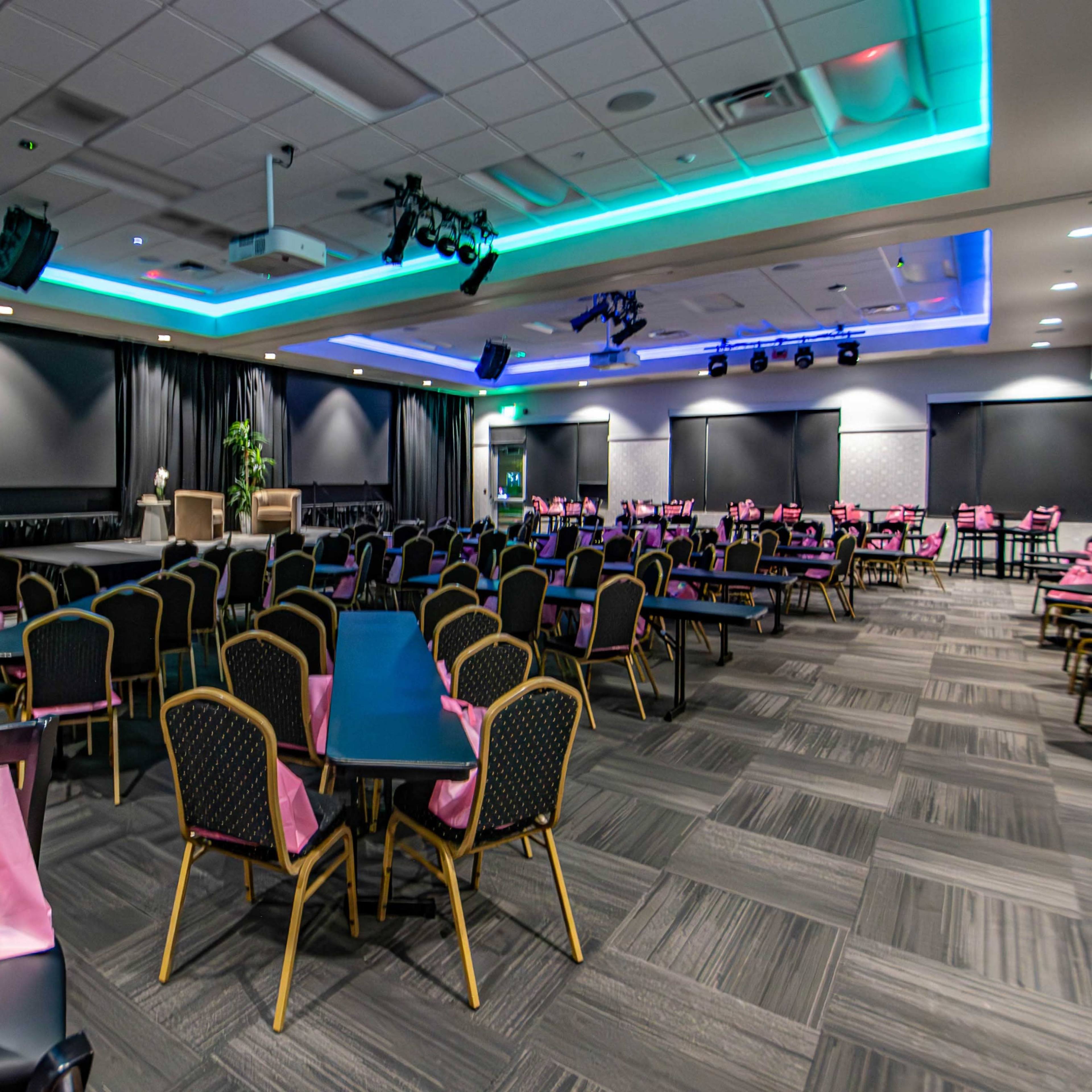 A spacious event room with black chairs and tables, decorated with pink bags on the tables, and equipped with audio-visual equipment and dim lighting.