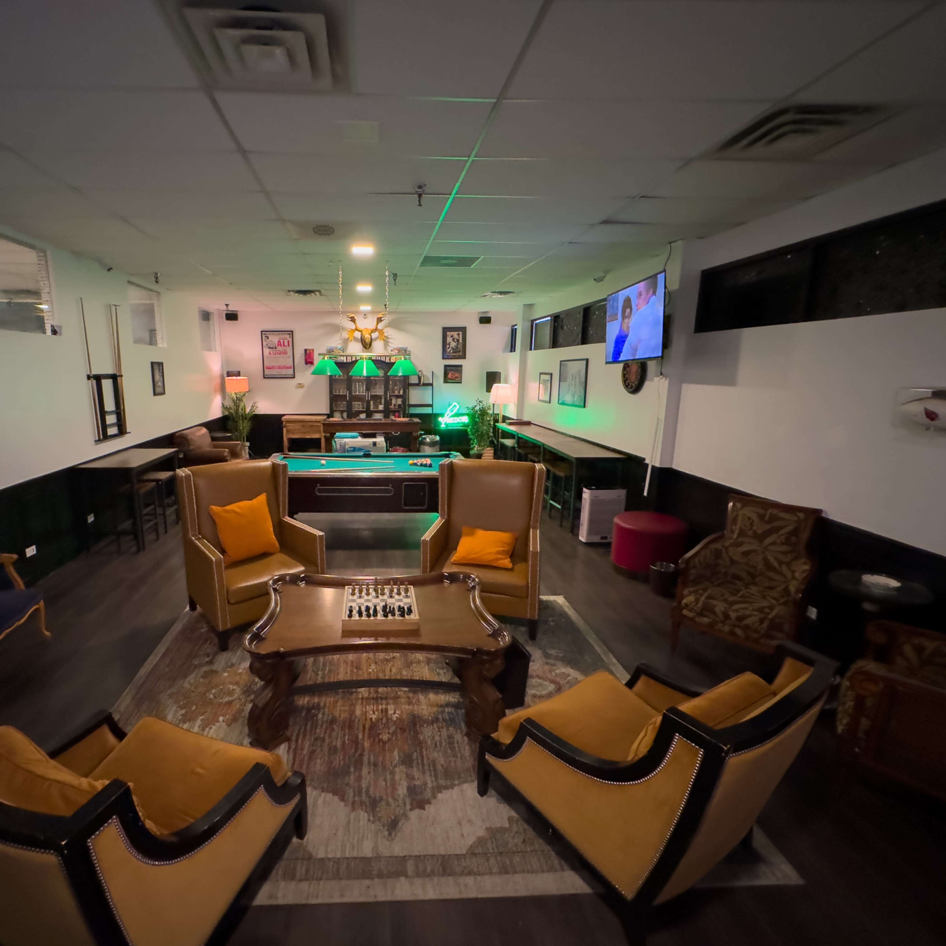 A lounge area features several armchairs and a coffee table on a patterned rug, with a bar and wall-mounted television in the background.