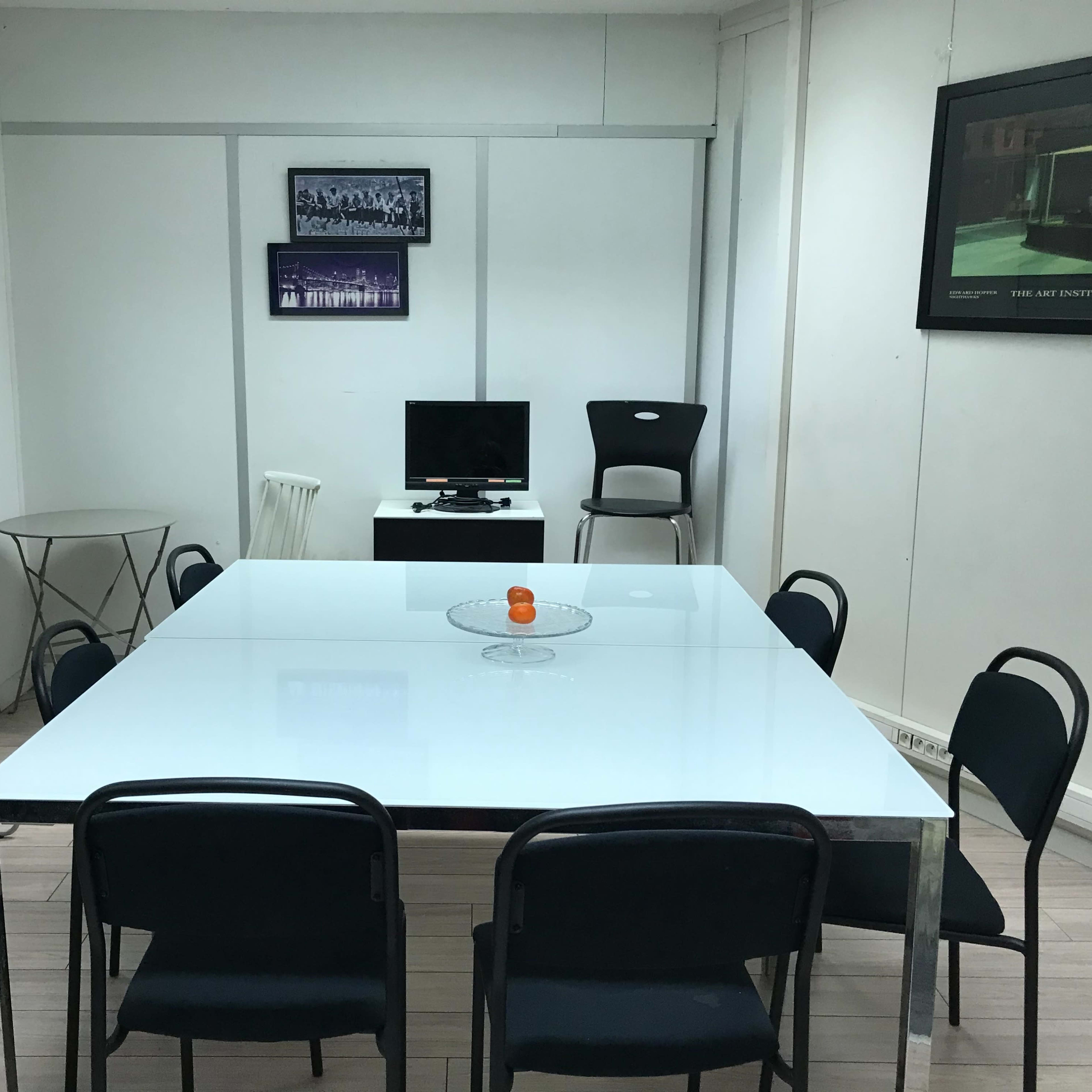 A conference room features a large white table surrounded by black chairs, with a red chair, a television, and framed artworks on the walls.