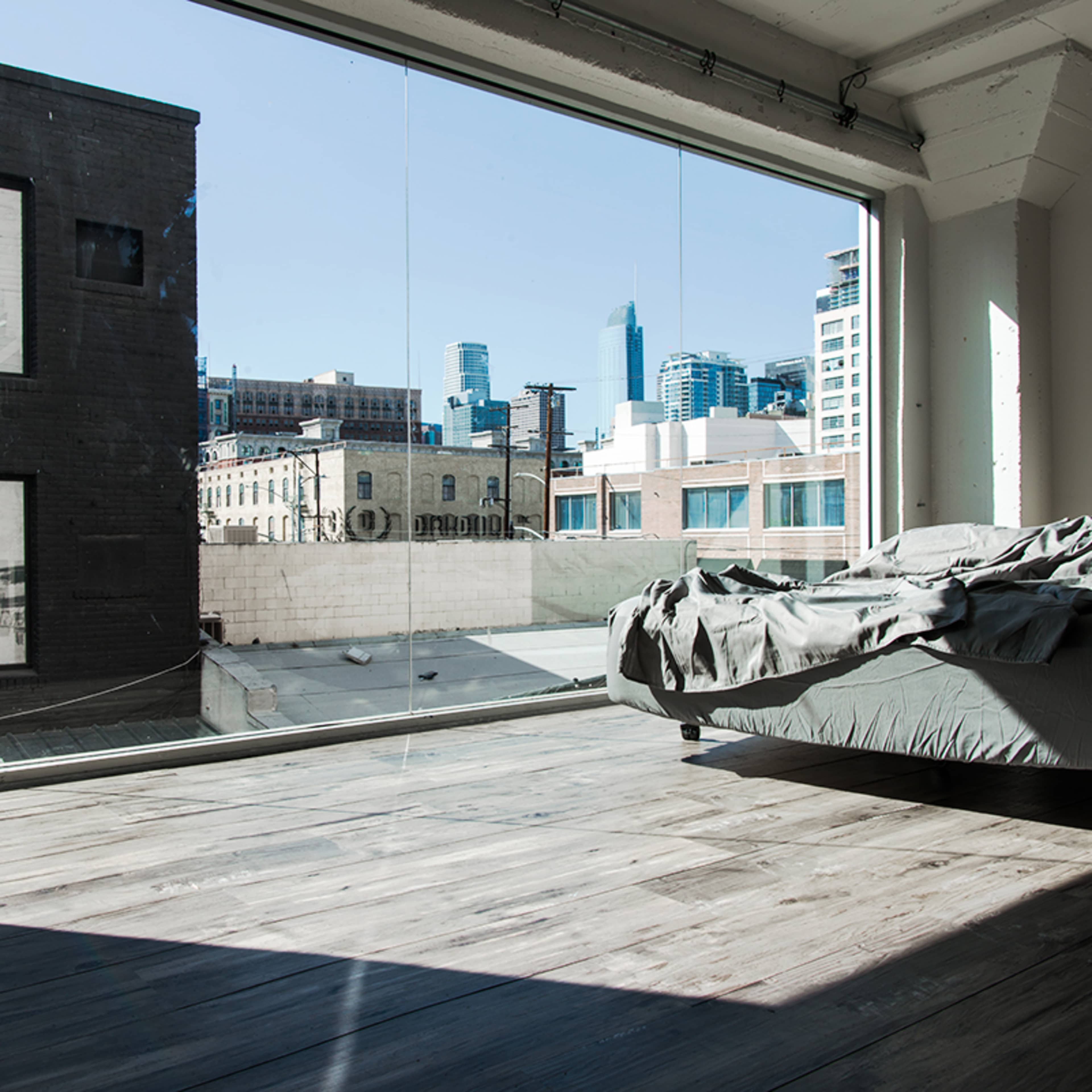 A sparse bedroom features a unmade bed against a large glass wall overlooking a cityscape.