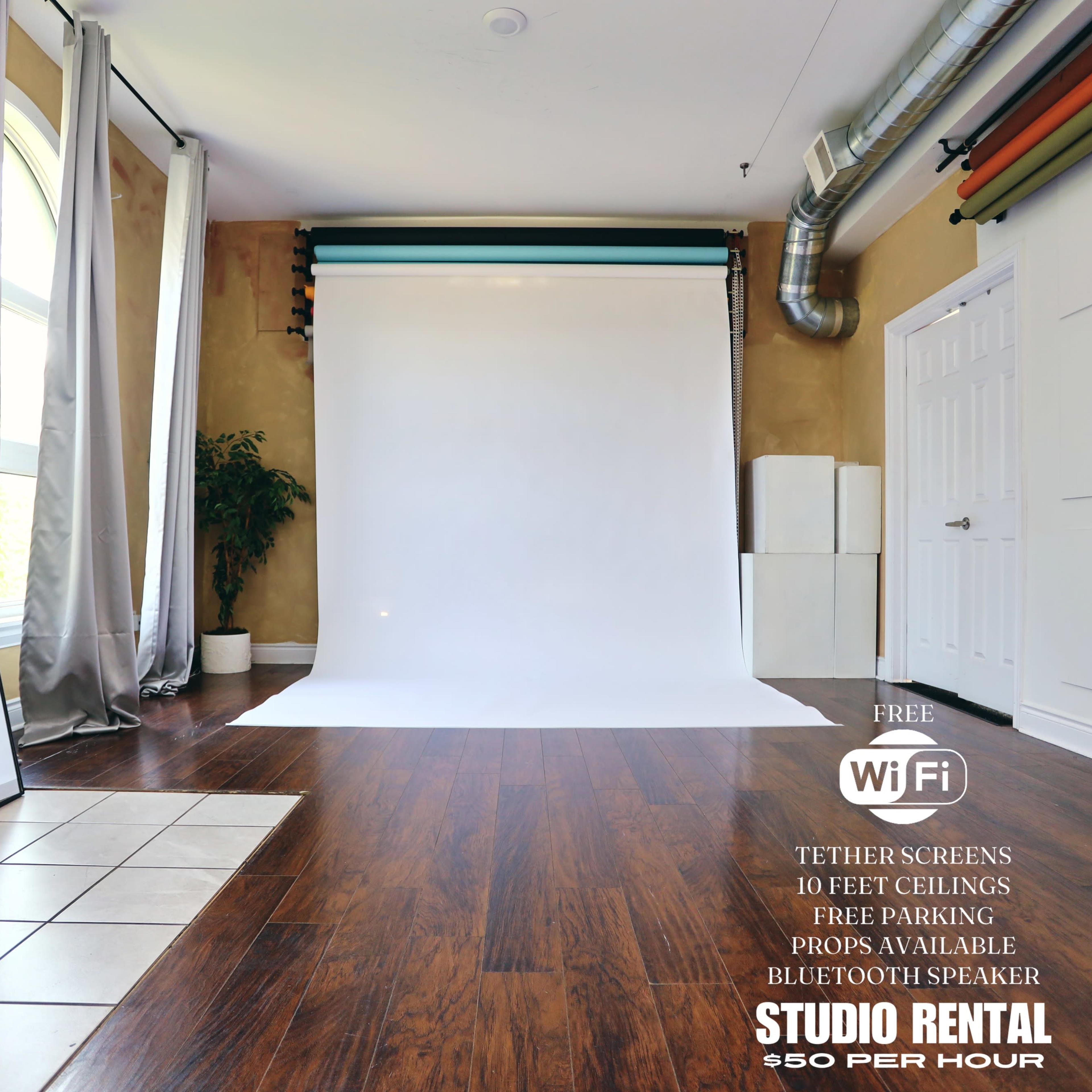 A spacious studio with a white backdrop, wooden floors, and various amenities including a Bluetooth speaker and WiFi, available for rental.