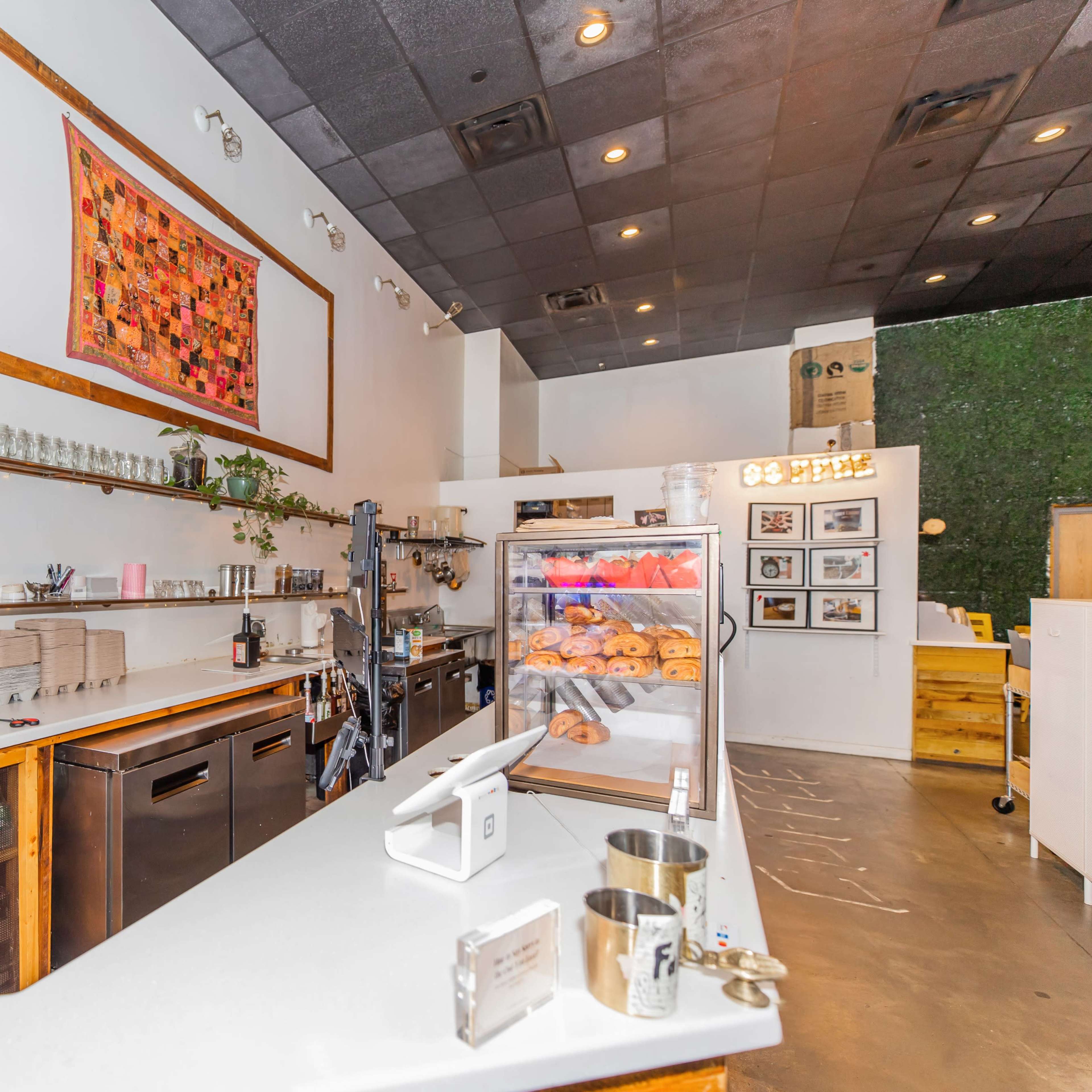 A modern café interior with a service counter, glass display case of pastries, and wooden shelving against a clean, bright wall.