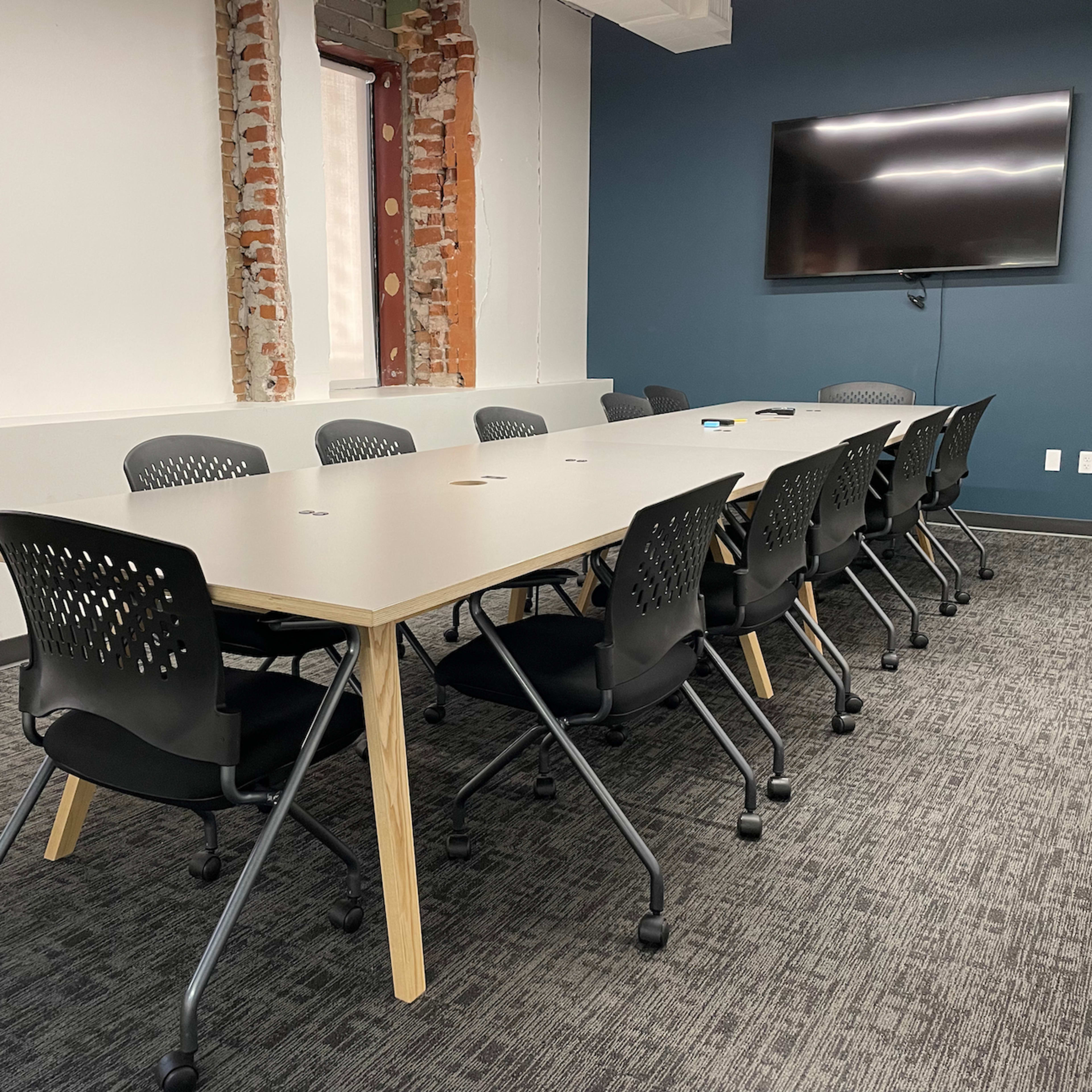 A long, rectangular table with black chairs is situated in a meeting room featuring exposed brick walls and a wall-mounted television.