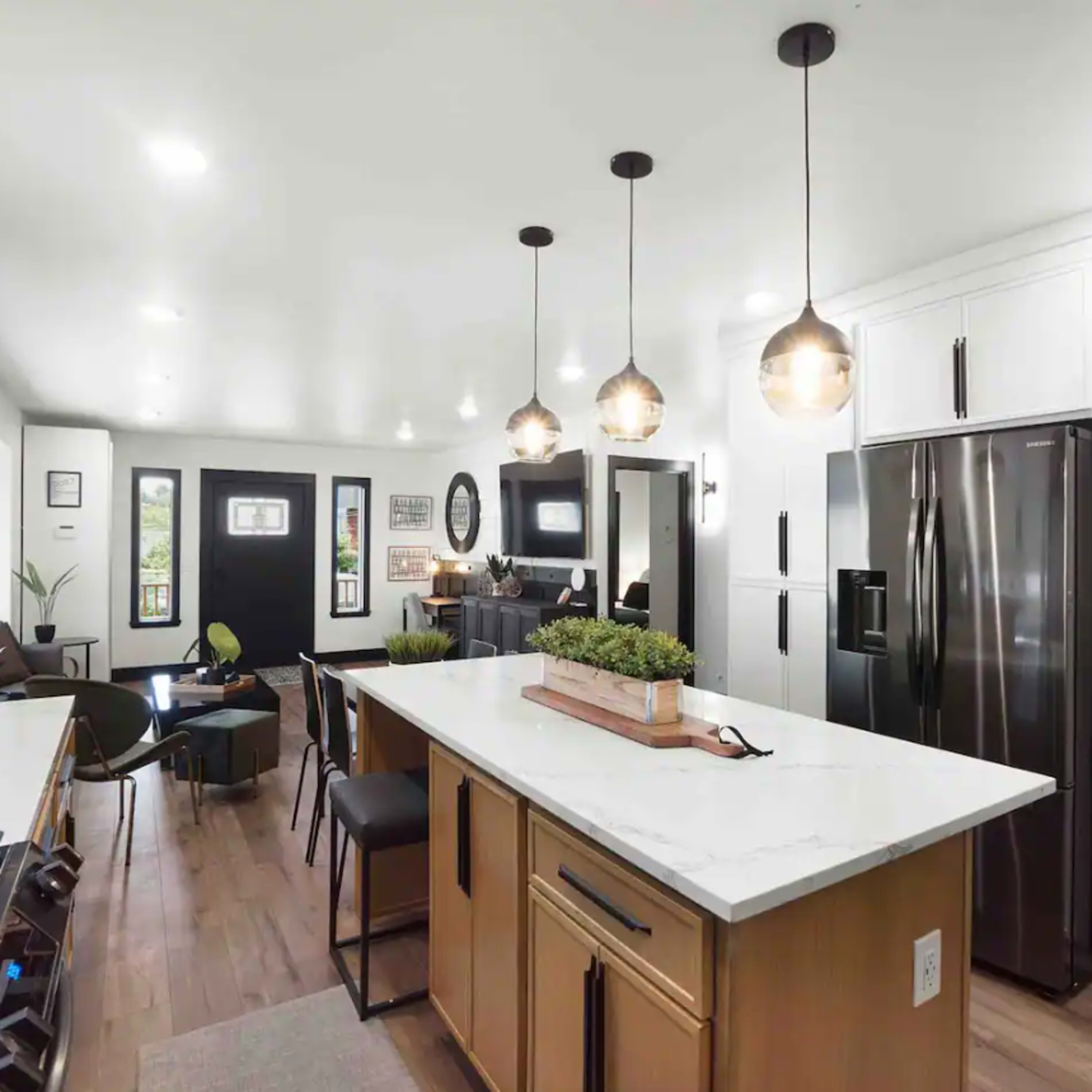 A modern kitchen features a large island with pendant lighting, stainless steel appliances, and a contemporary design with white cabinetry and wooden accents.