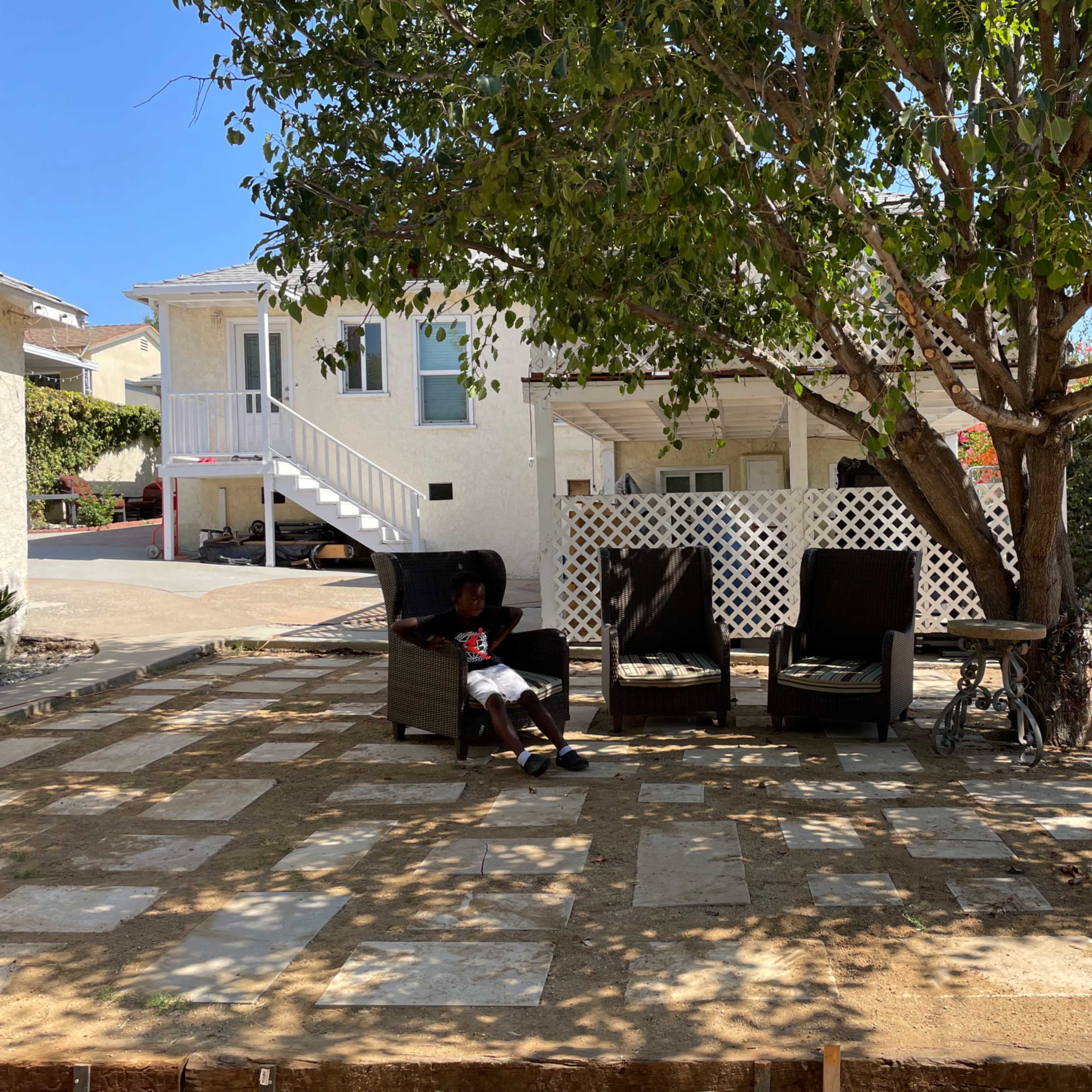 A person sits on a chair under a tree in a patio area with stone paving and a residential house in the background.