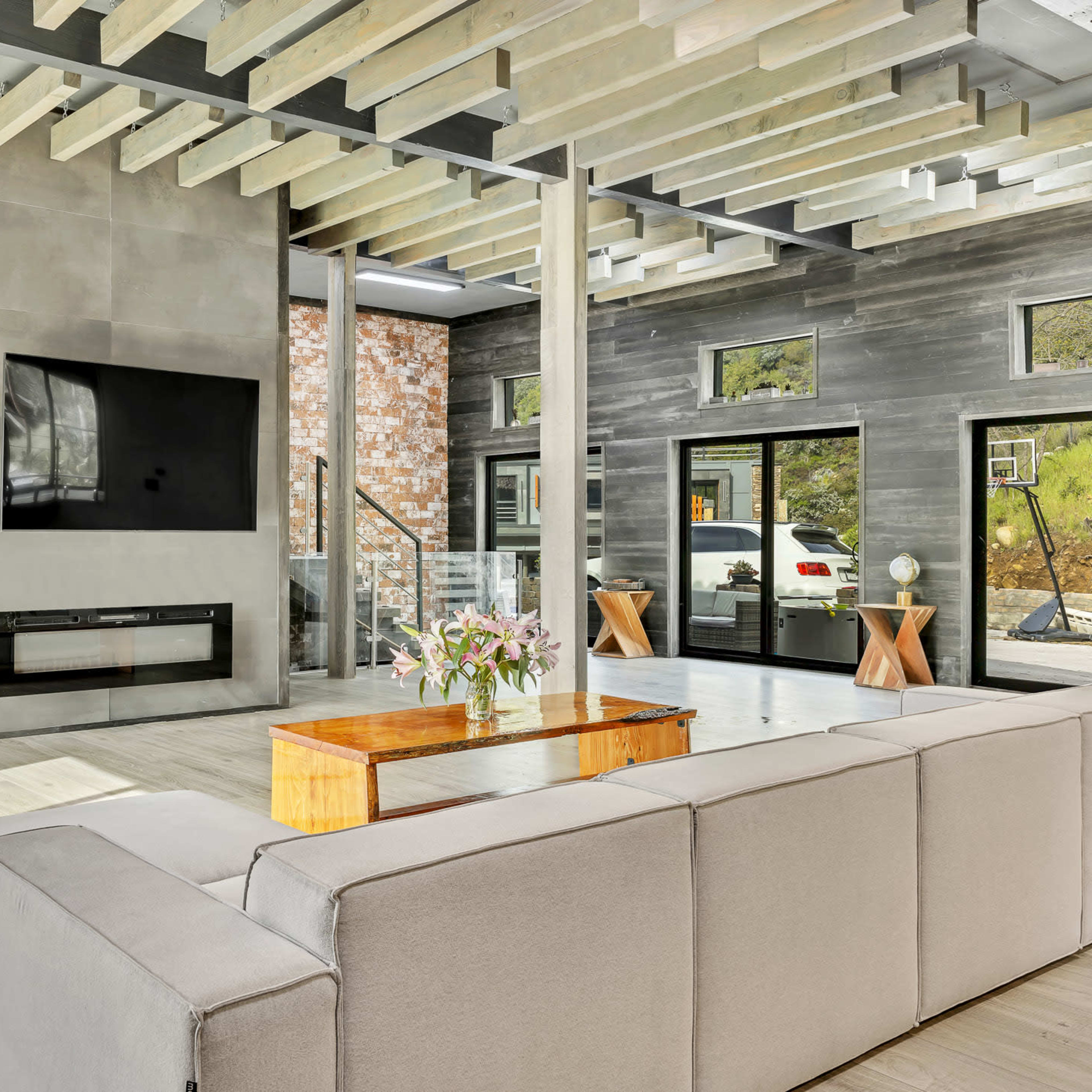 A modern living room with a large window, minimalist furniture, a flat-screen TV mounted on a concrete wall, and natural light streaming in from multiple angles.