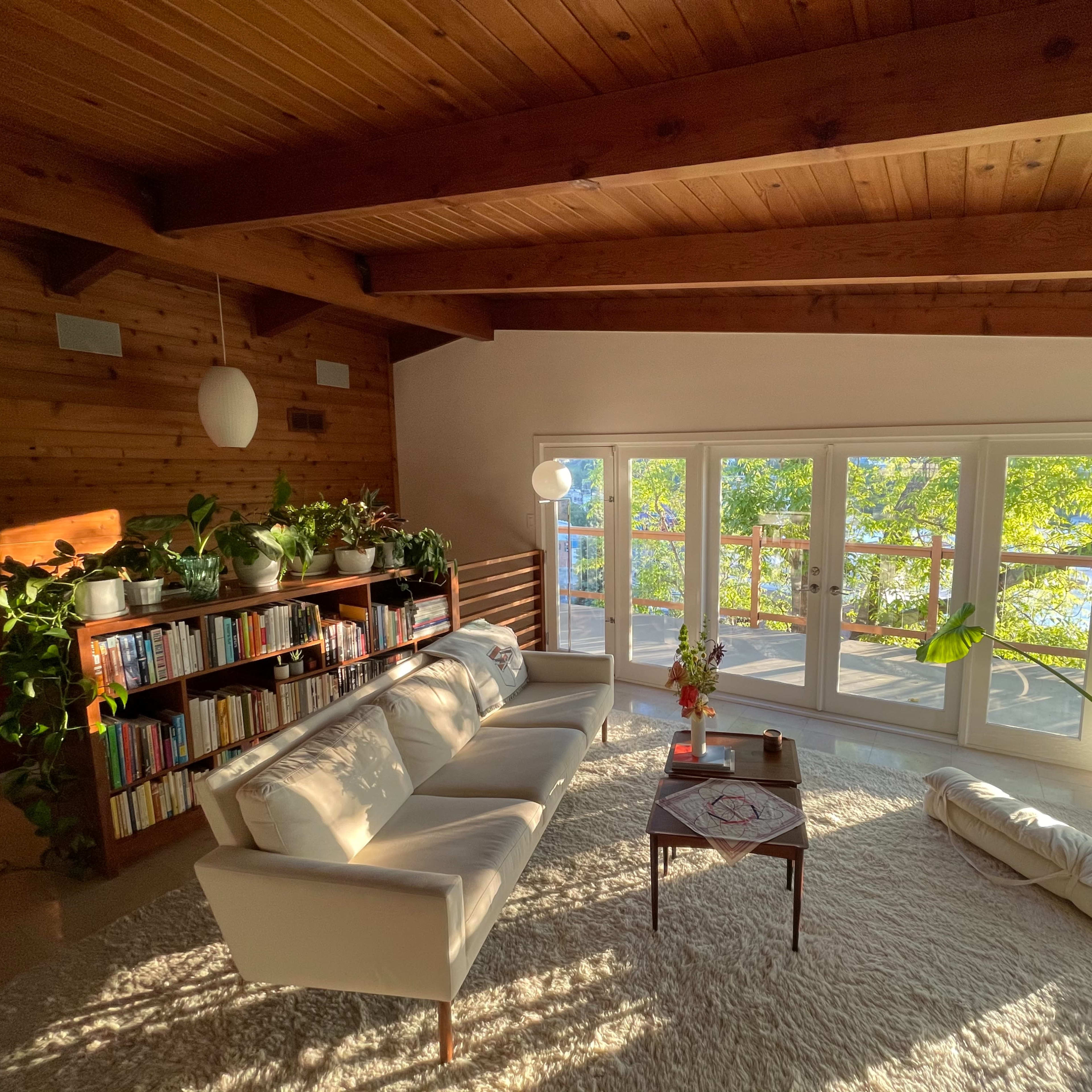 A cozy living room features a white sofa, a coffee table, a bookshelf filled with books, and large windows overlooking a green landscape.