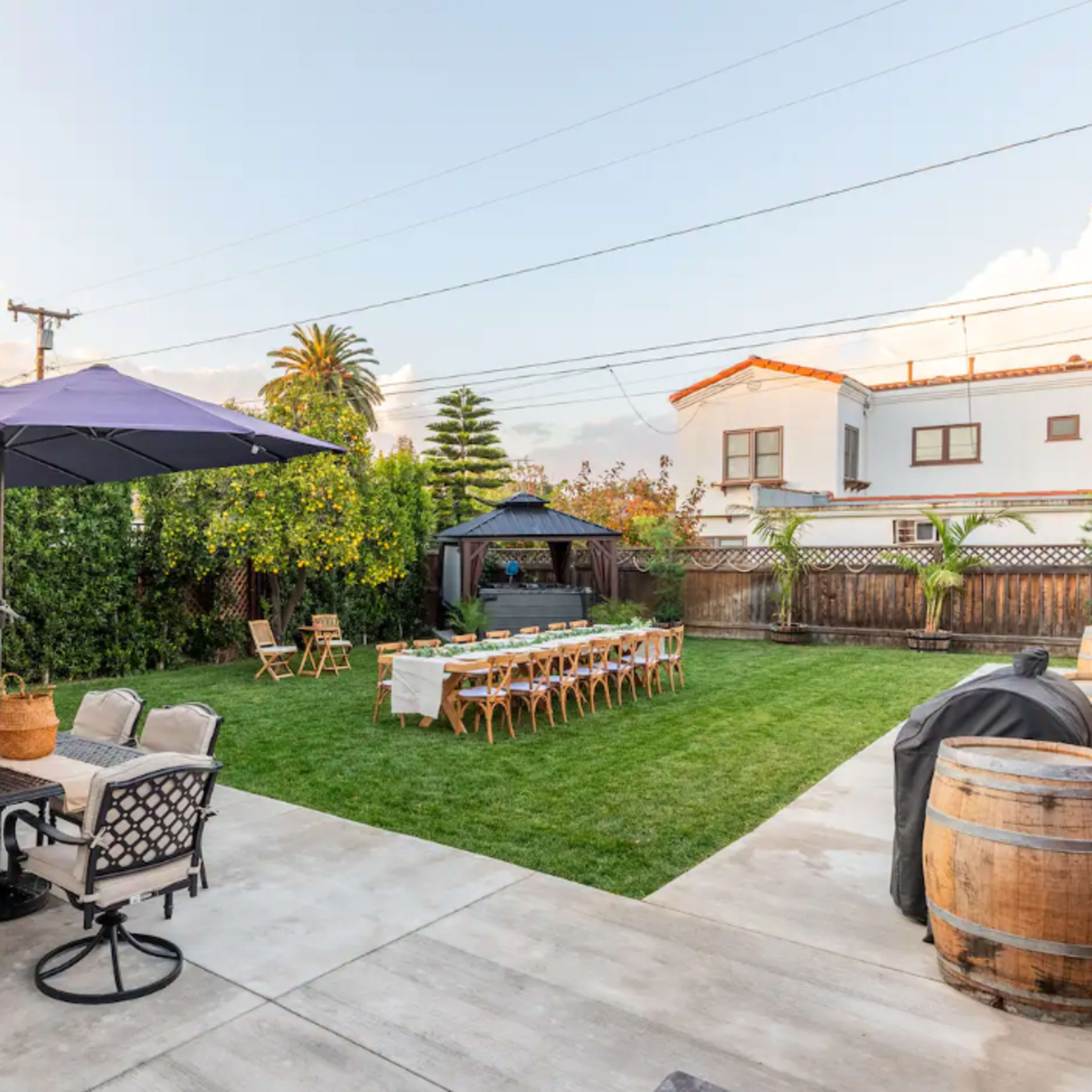 A spacious outdoor area featuring a dining setup with a long table and chairs, shaded by an umbrella, alongside a seating arrangement with a table and chairs, all surrounded by greenery.