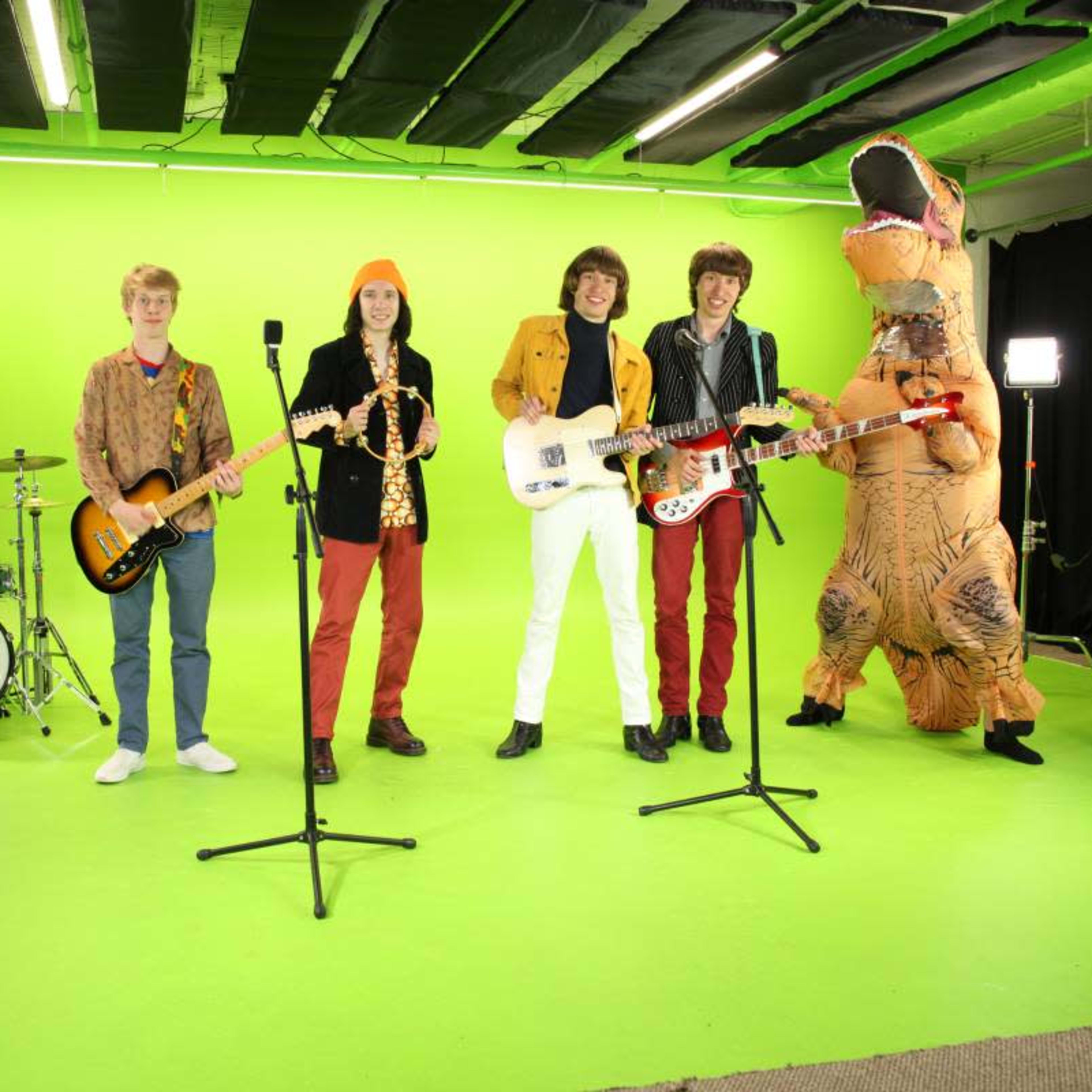 A band of five musicians in colorful outfits poses with their instruments on a bright green studio set, while one member in a dinosaur costume stands among them.