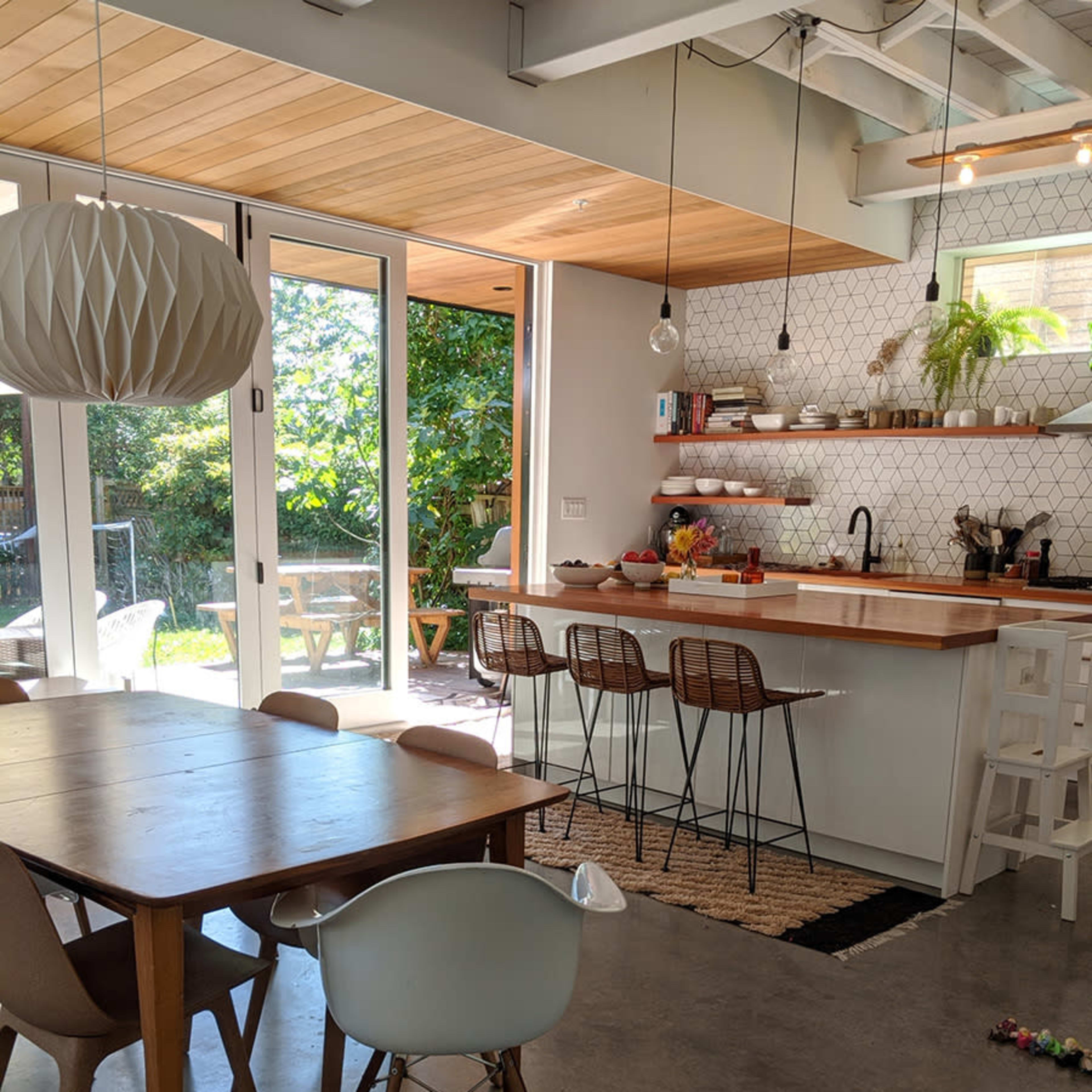 A modern kitchen and dining area features a wooden table, bentwood chairs, a long counter with stools, and large windows offering a view of the garden.