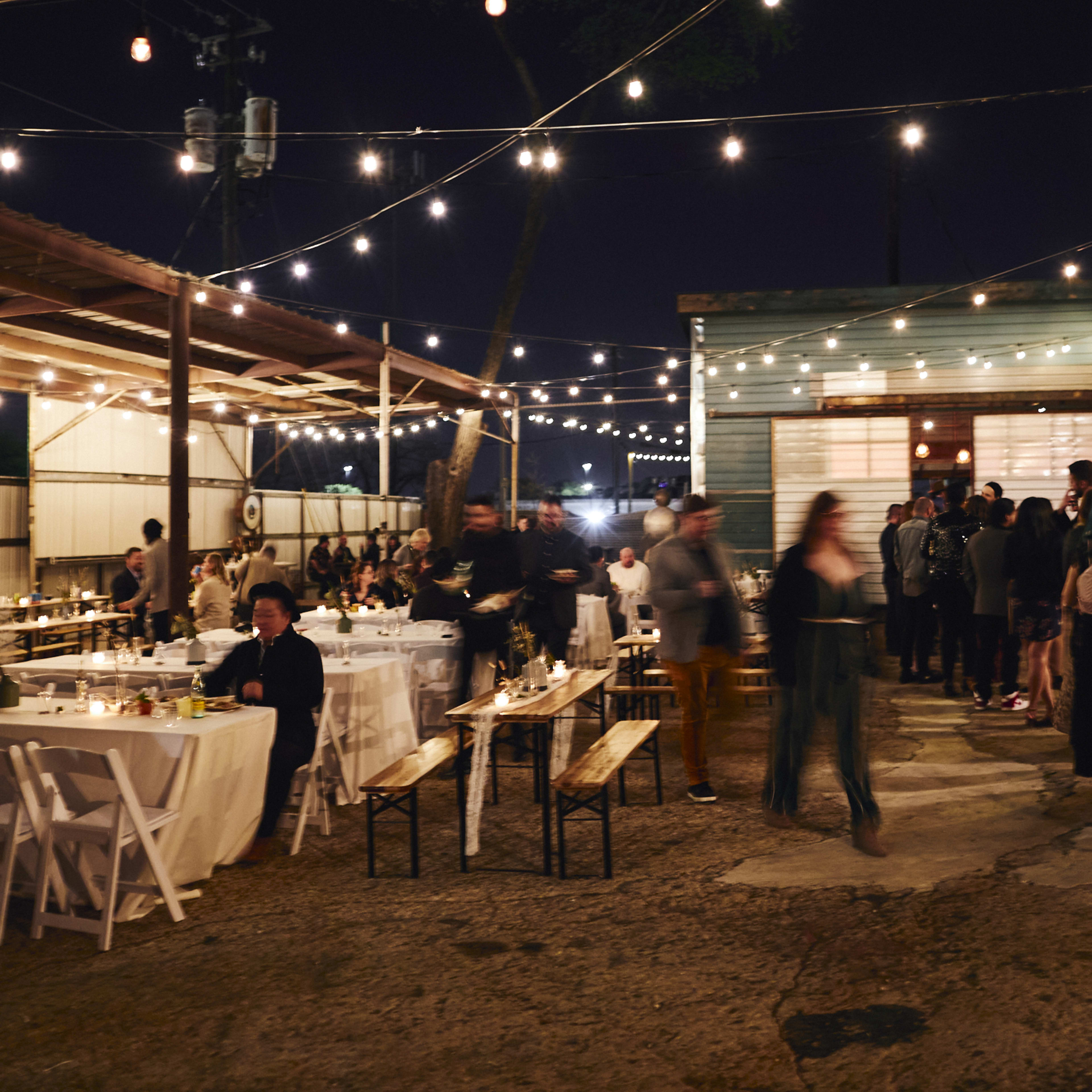 A lively outdoor gathering takes place at night, with guests seated at long tables under string lights, while others form a line near a building.