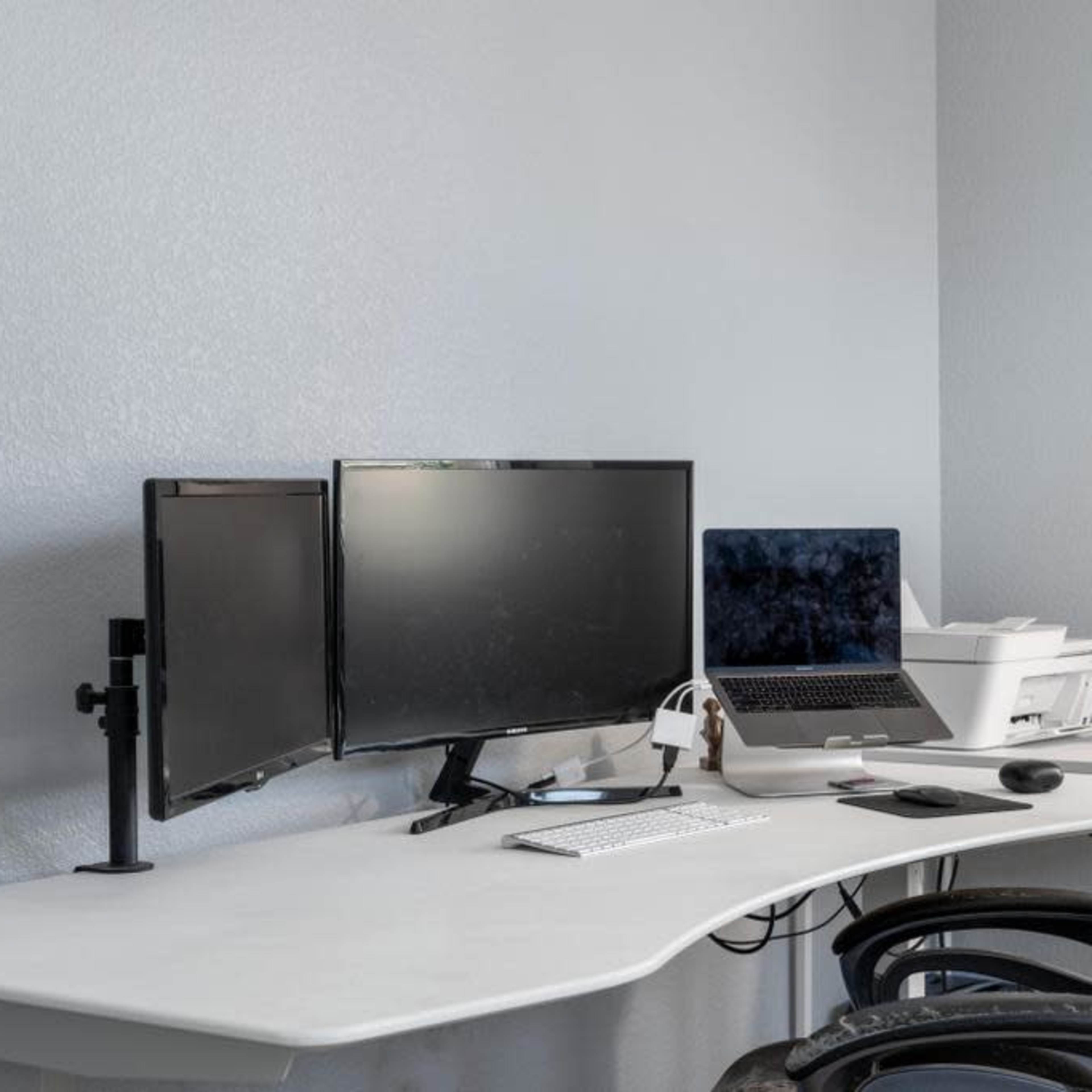 A modern office setup features two monitors, a laptop, a printer, and a chair at a white desk illuminated by a floor lamp.