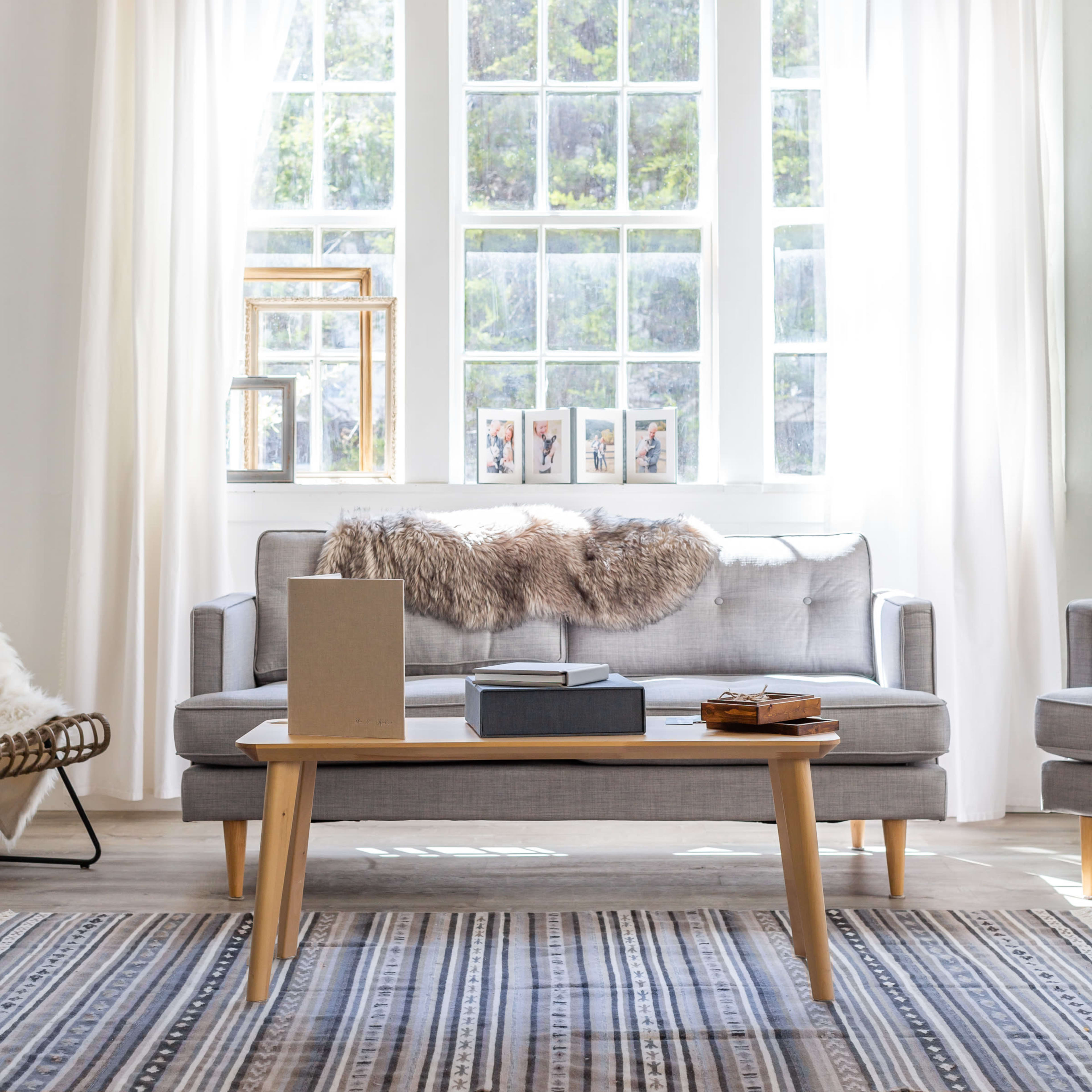 A light-filled living room features a gray sofa with a fur throw, a wooden coffee table, and large windows with sheer curtains.