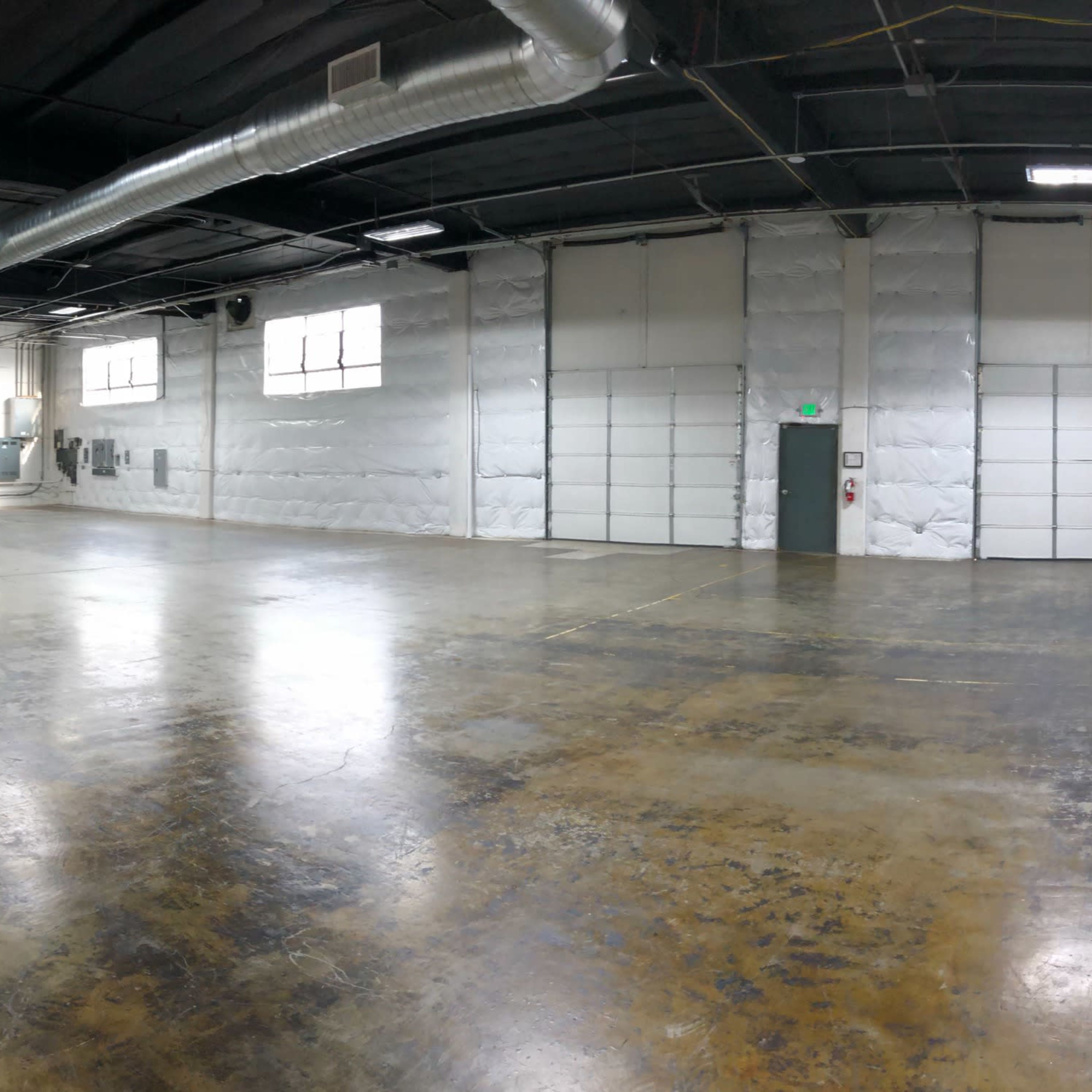 A large, empty industrial warehouse space with concrete flooring and metal overhead lighting.