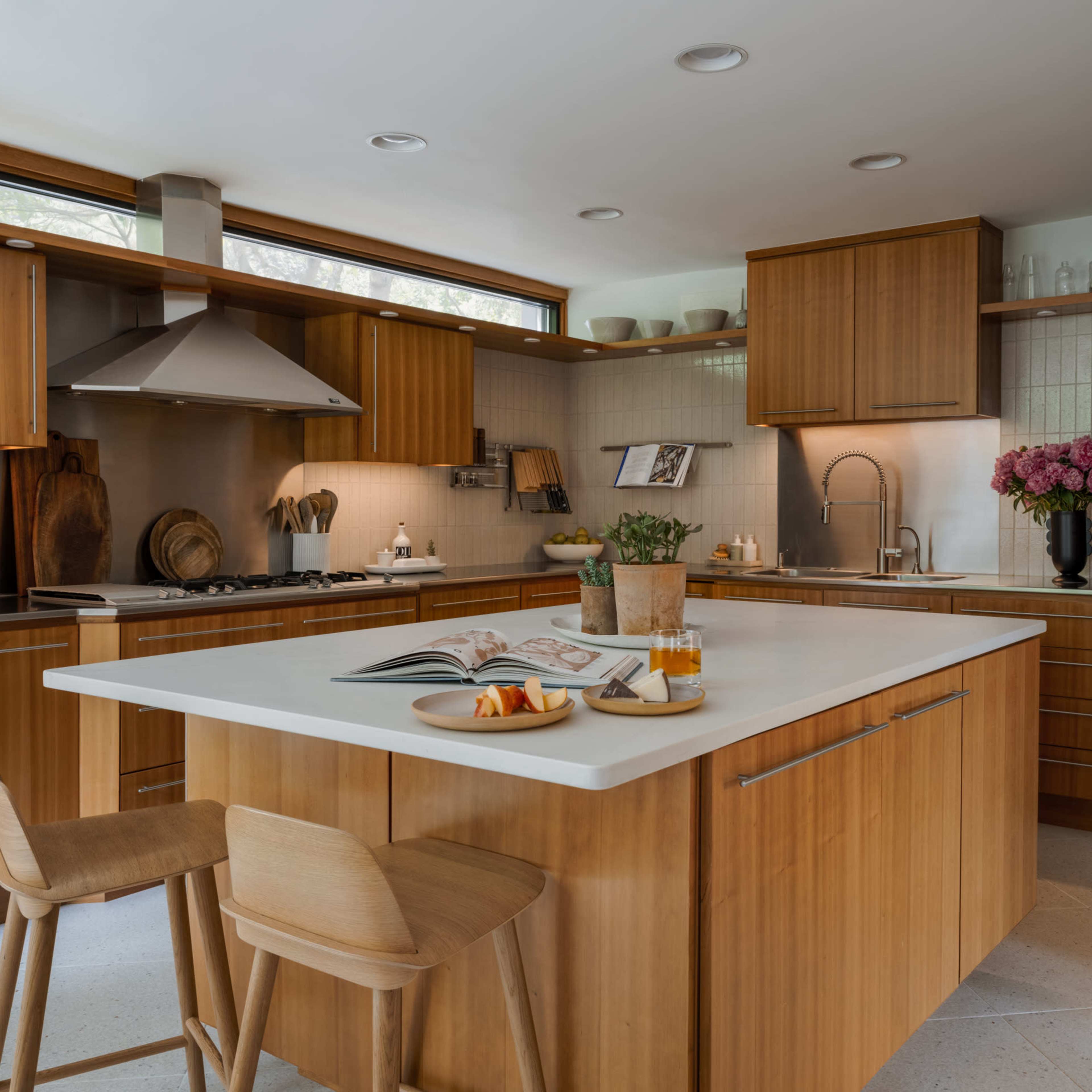 A modern kitchen features wooden cabinetry, a central island with bar stools, and a collection of cooking utensils and fresh flowers on the countertop.
