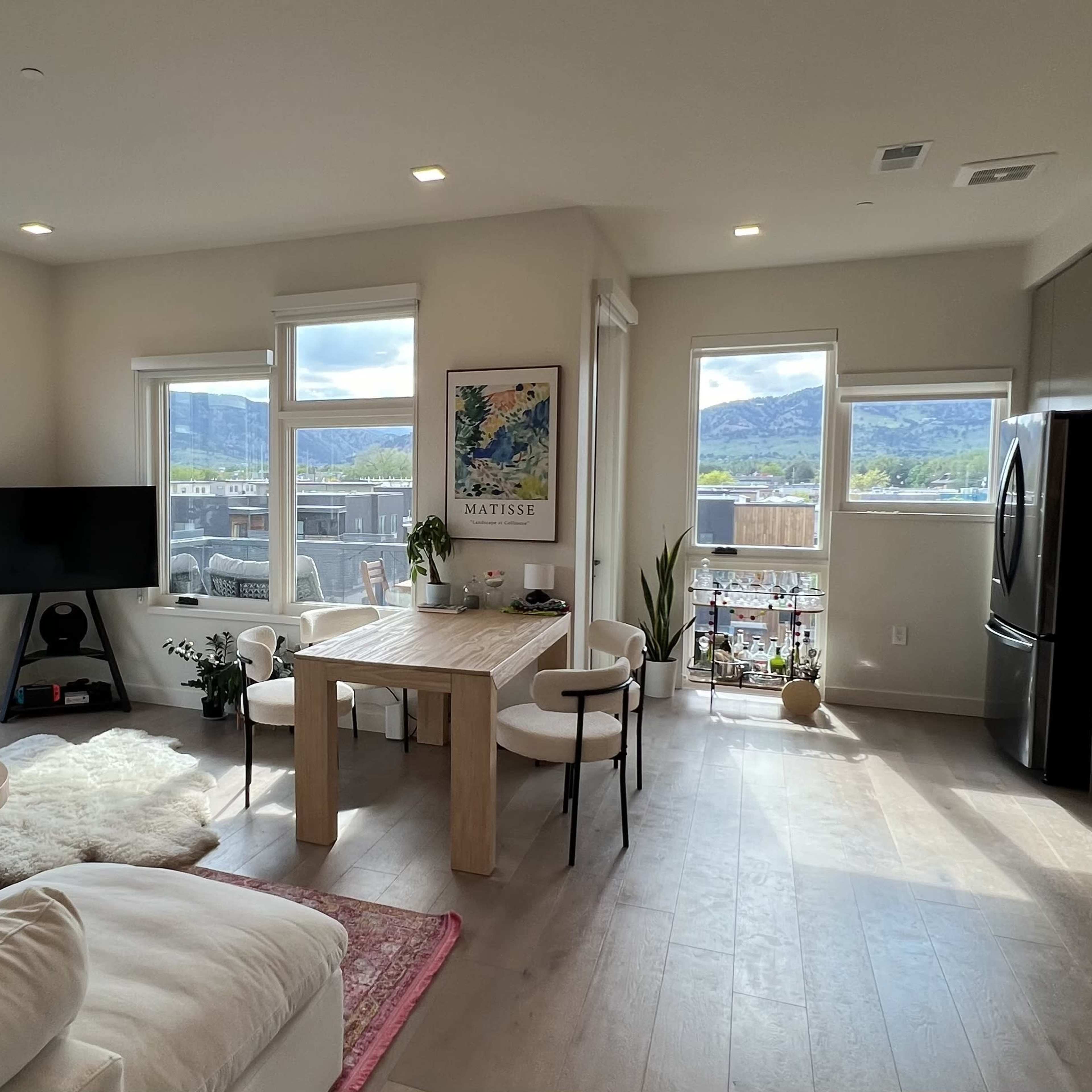A modern living room and dining area with large windows showcasing a mountain view, featuring a white sofa, a wooden dining table, and a television.