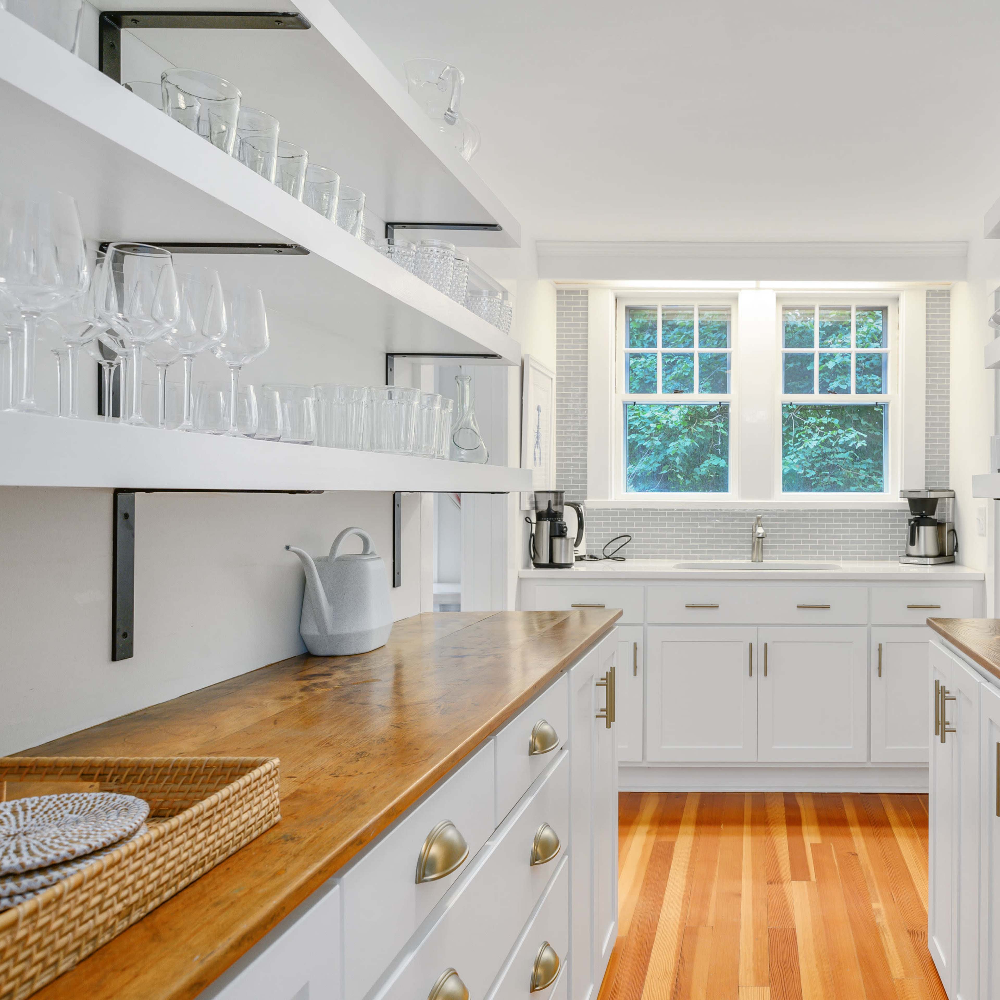 A bright pantry features white shelves lined with glassware and dishware, flanking wooden countertops and a window above a sink.