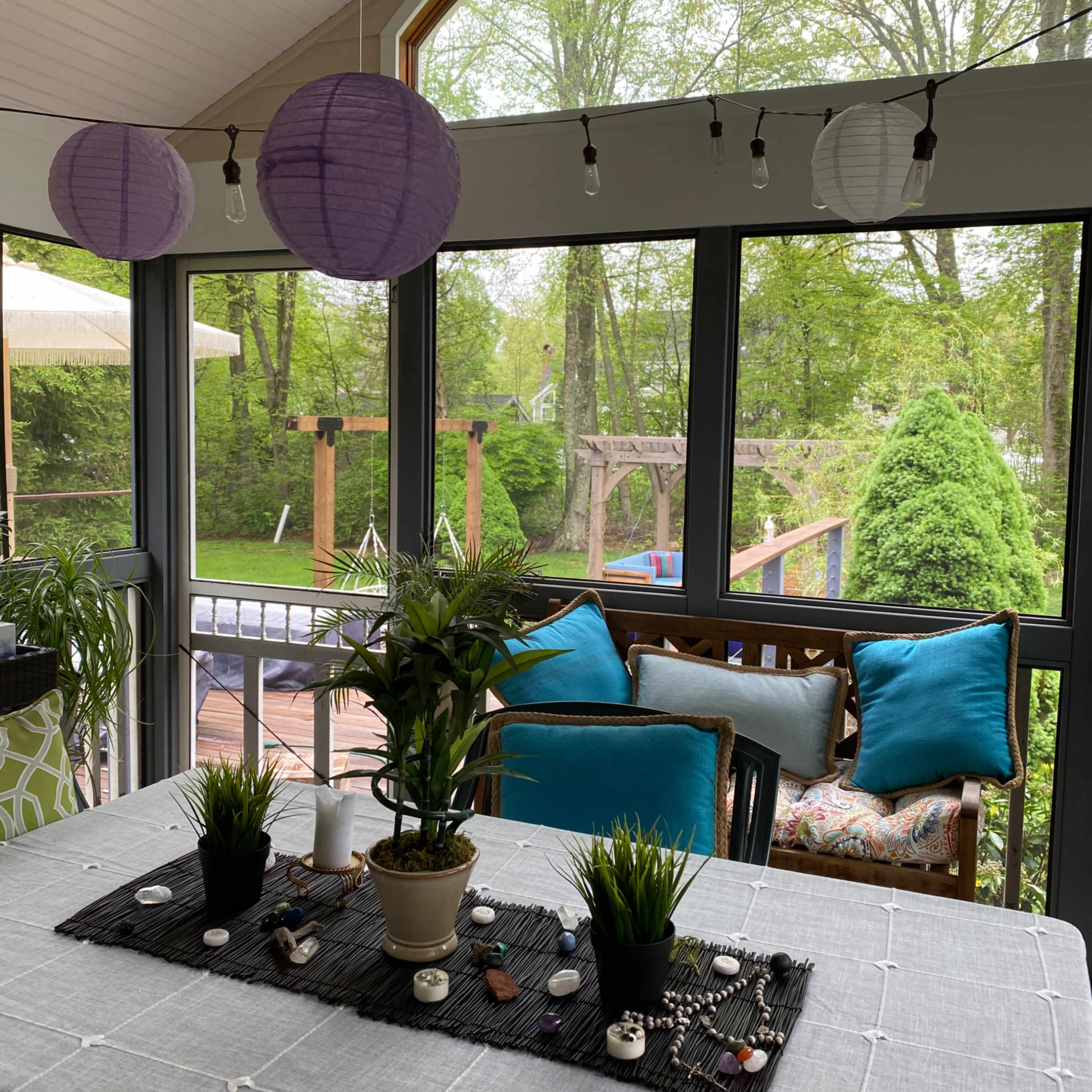 A bright sunroom features a dining table adorned with plants and decorative stones, surrounded by large windows overlooking a green outdoor space.