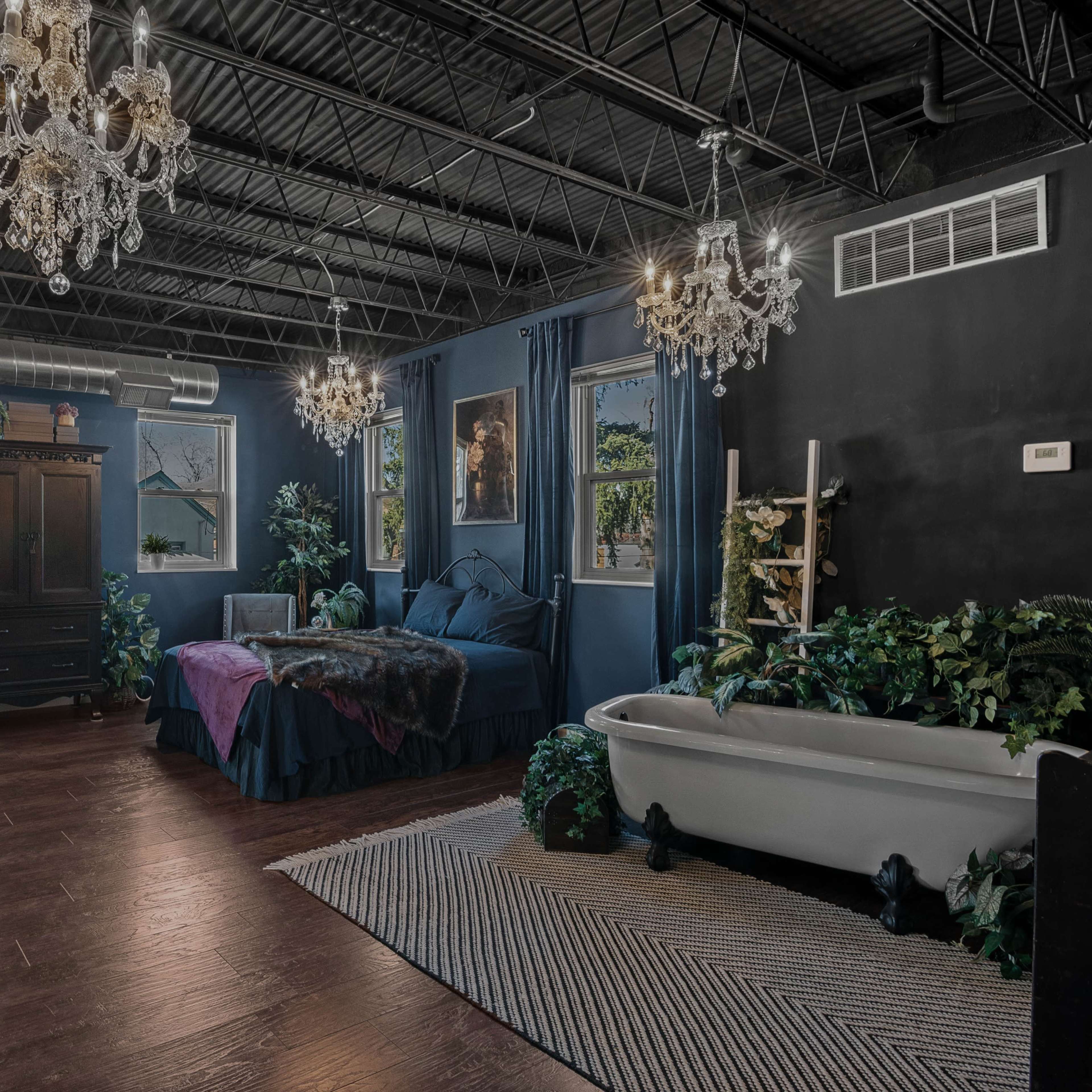 A spacious bedroom featuring two chandeliers, a dark color scheme, a large bed, a vintage bathtub, and various plants along the walls.