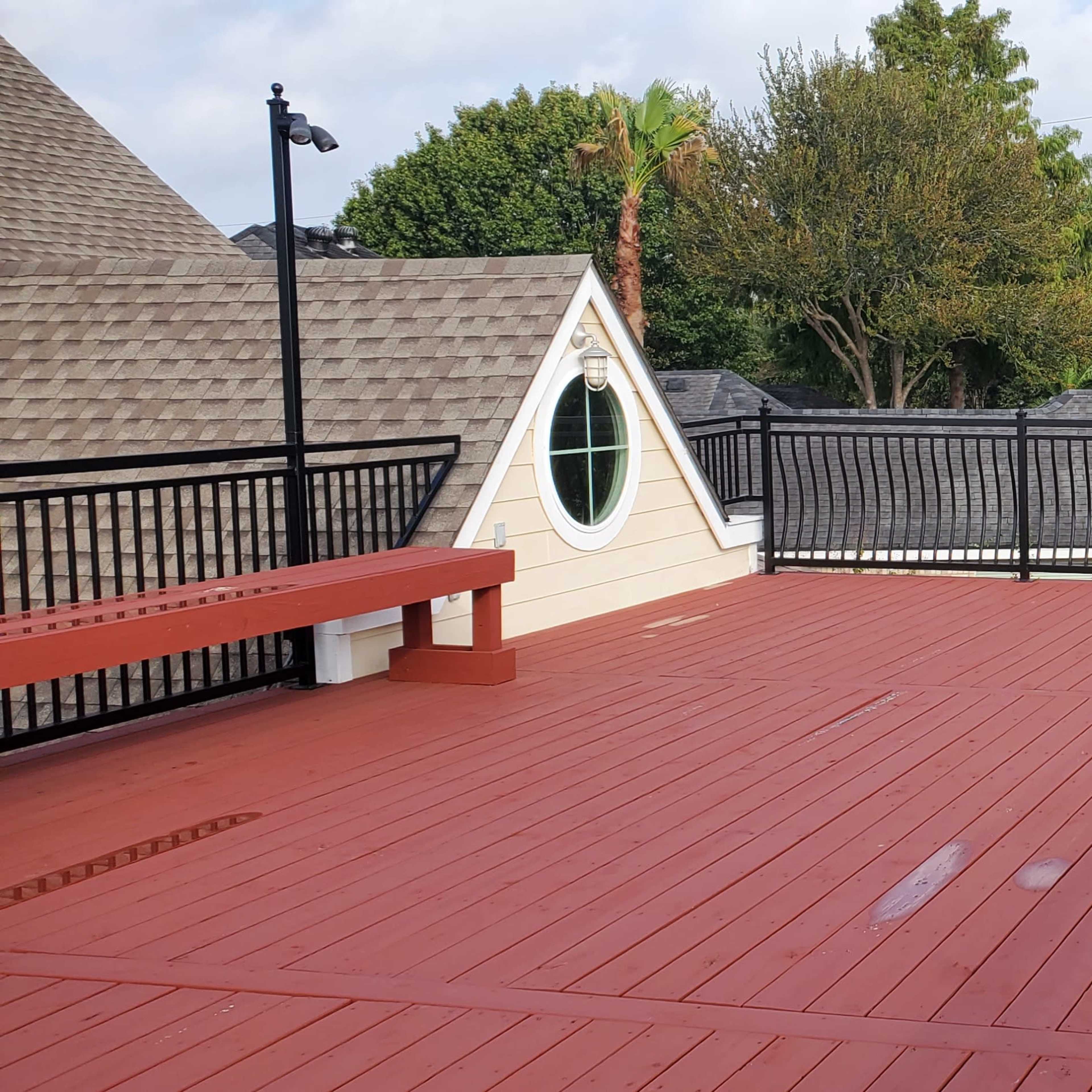A red wooden deck with a black railing, adjacent to a house and surrounded by trees and greenery.