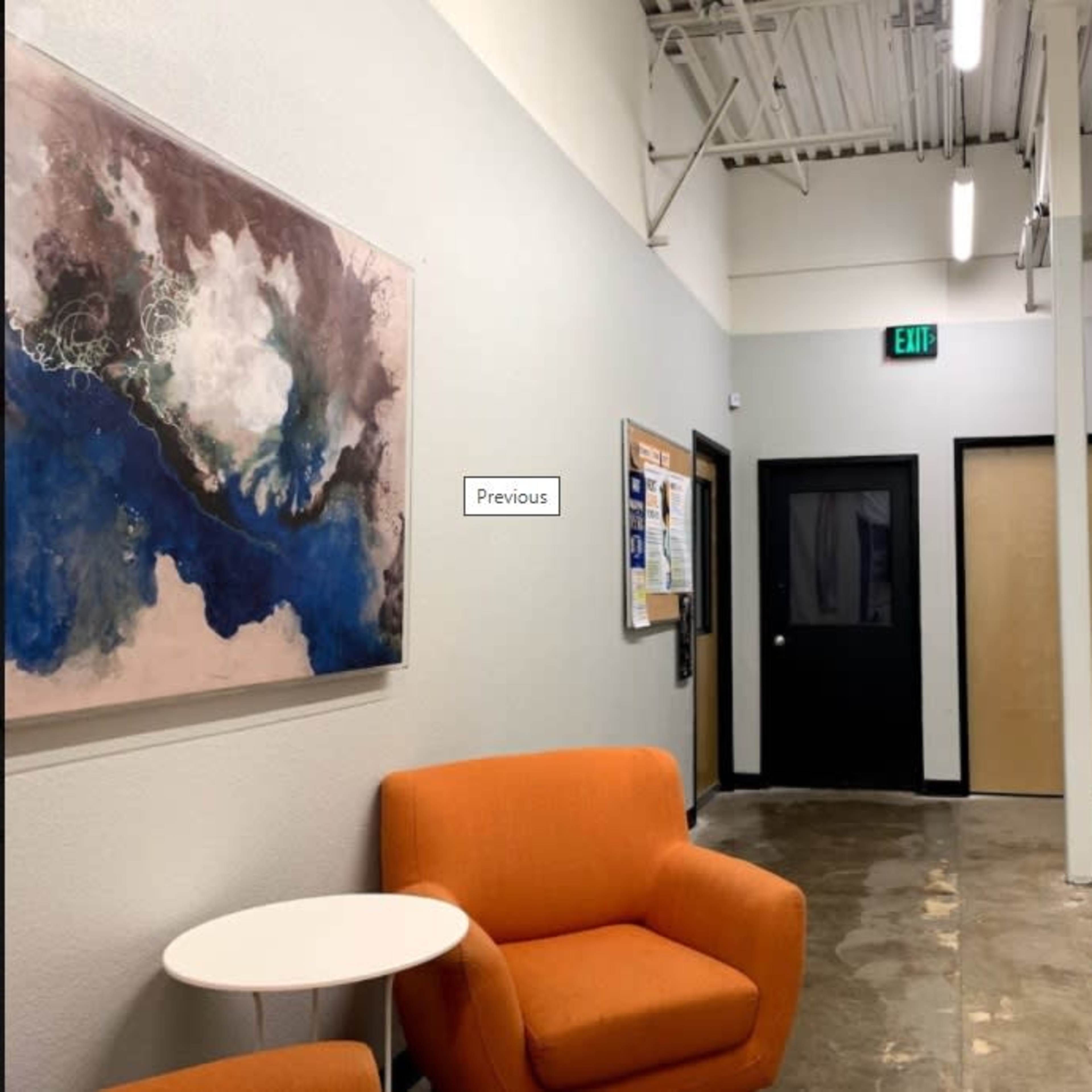 A hallway with orange chairs, a small white table, a large artwork on the wall, and doors leading to other rooms.
