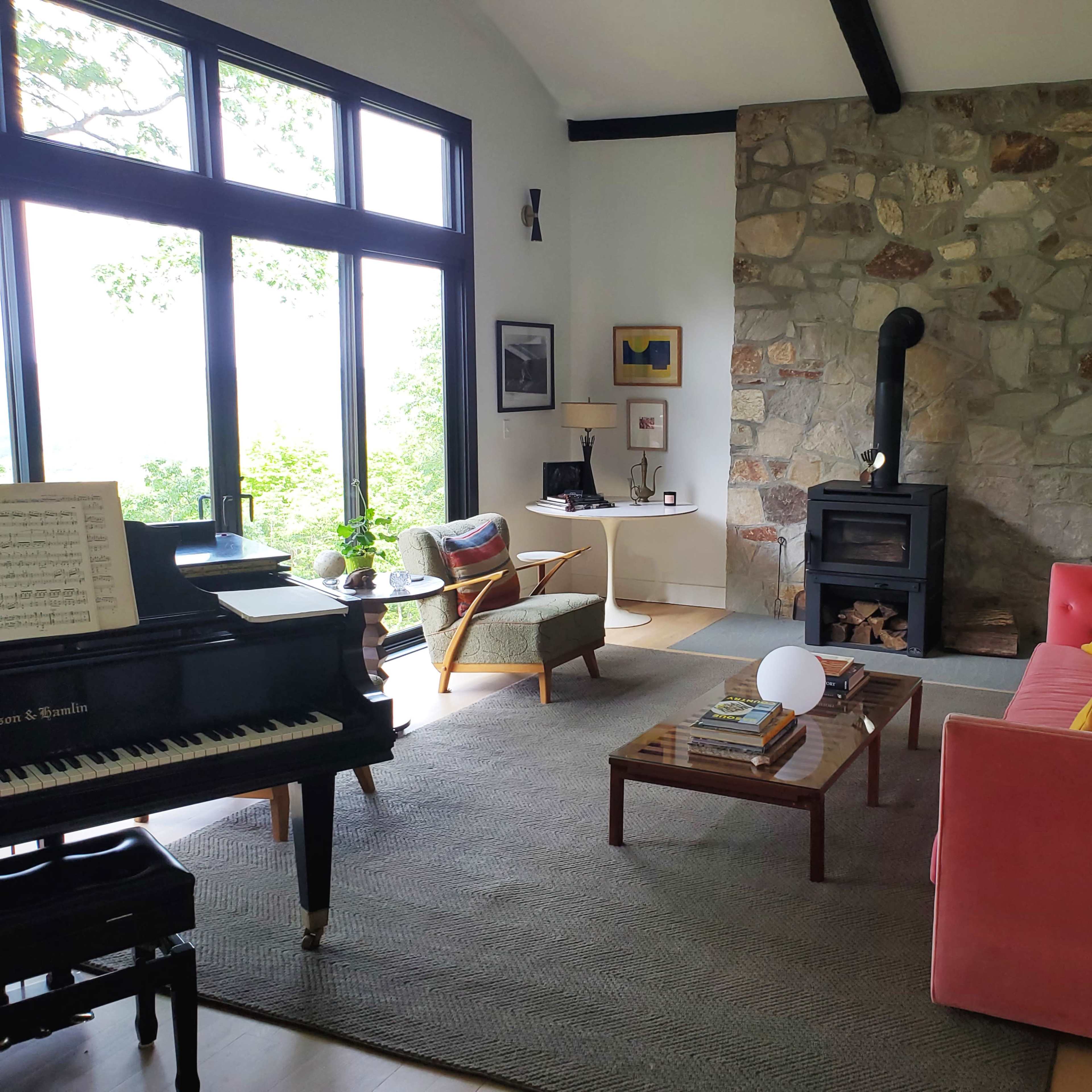 A cozy living room with a pink sofa, a piano, a stone fireplace, and large windows allowing natural light.