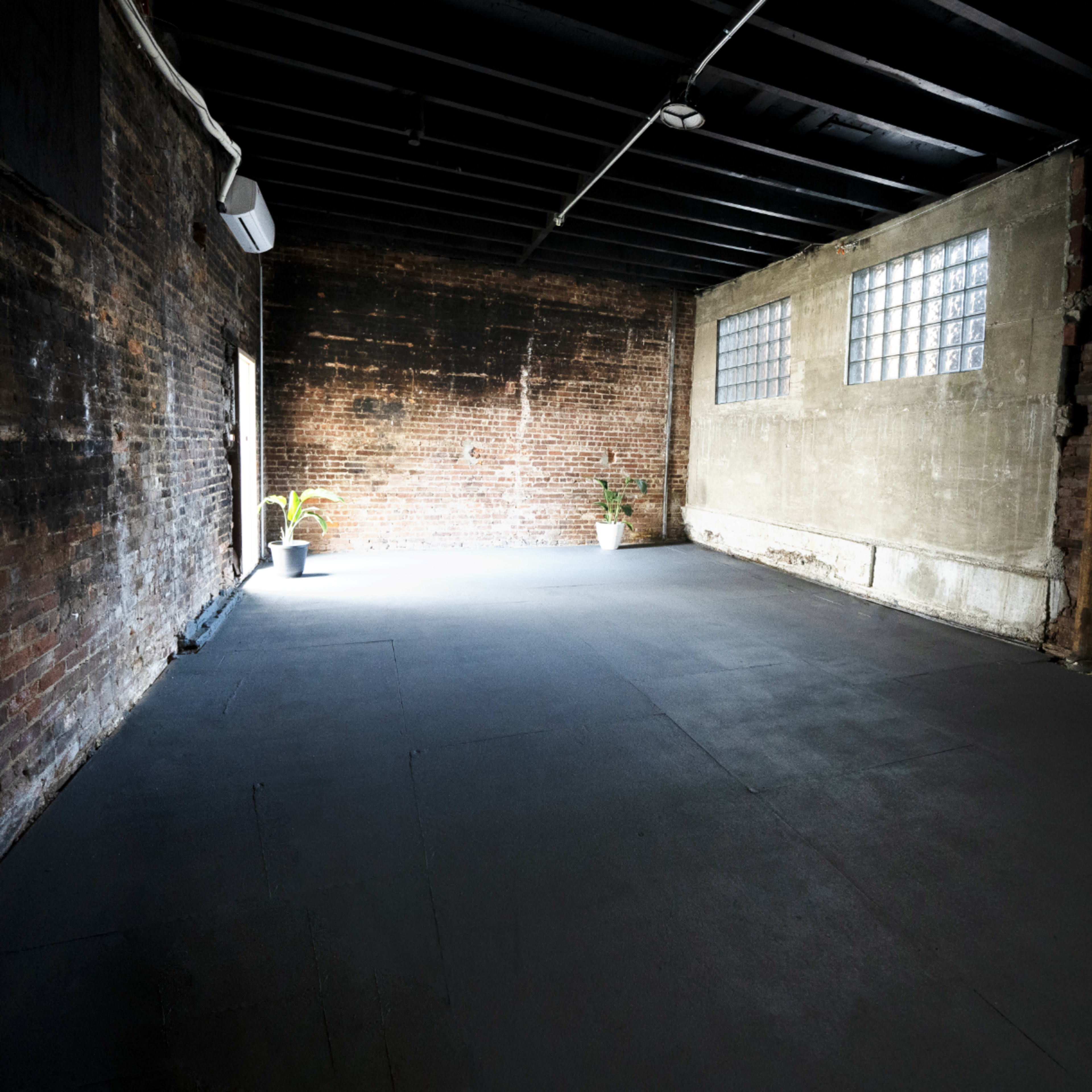An empty industrial room with exposed brick walls and a concrete floor, featuring a single potted plant in one corner.