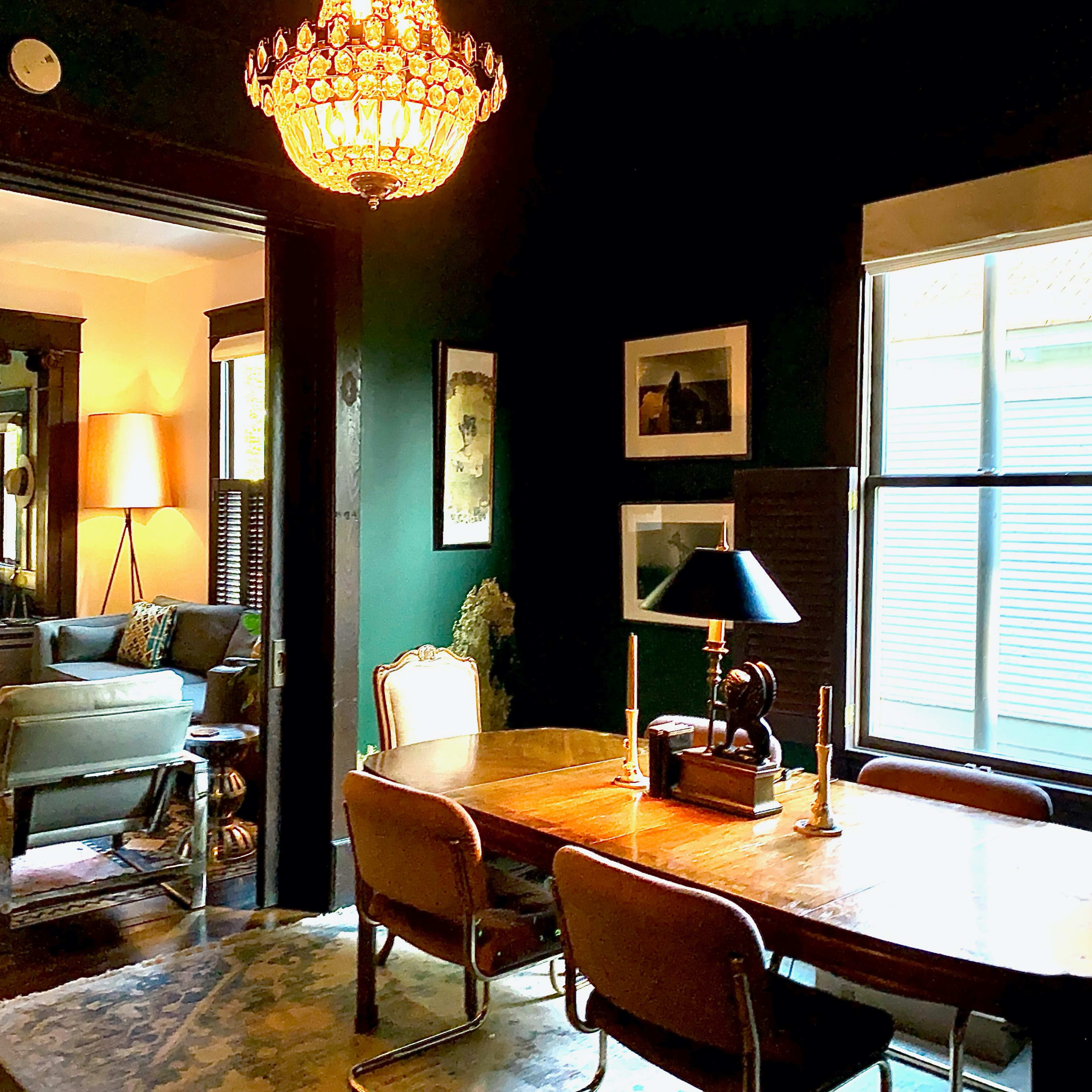 A dining area features a wooden table with chairs, a lamp, and a chandelier, while a living room with a guitar and plant is visible through an open doorway.