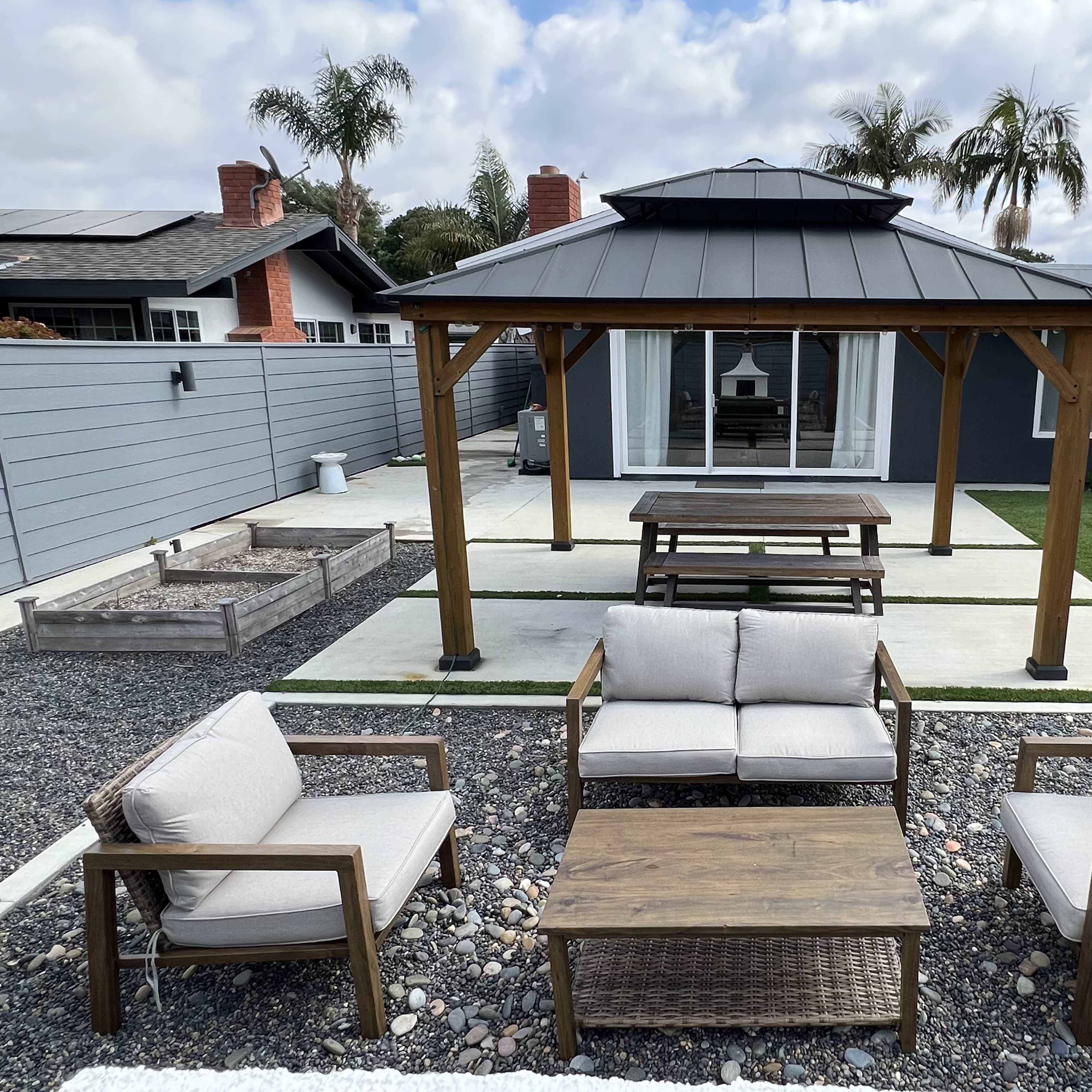 A backyard with a gazebo, a rectangular wooden table, and four cushioned seating chairs arranged around a stone patio area.