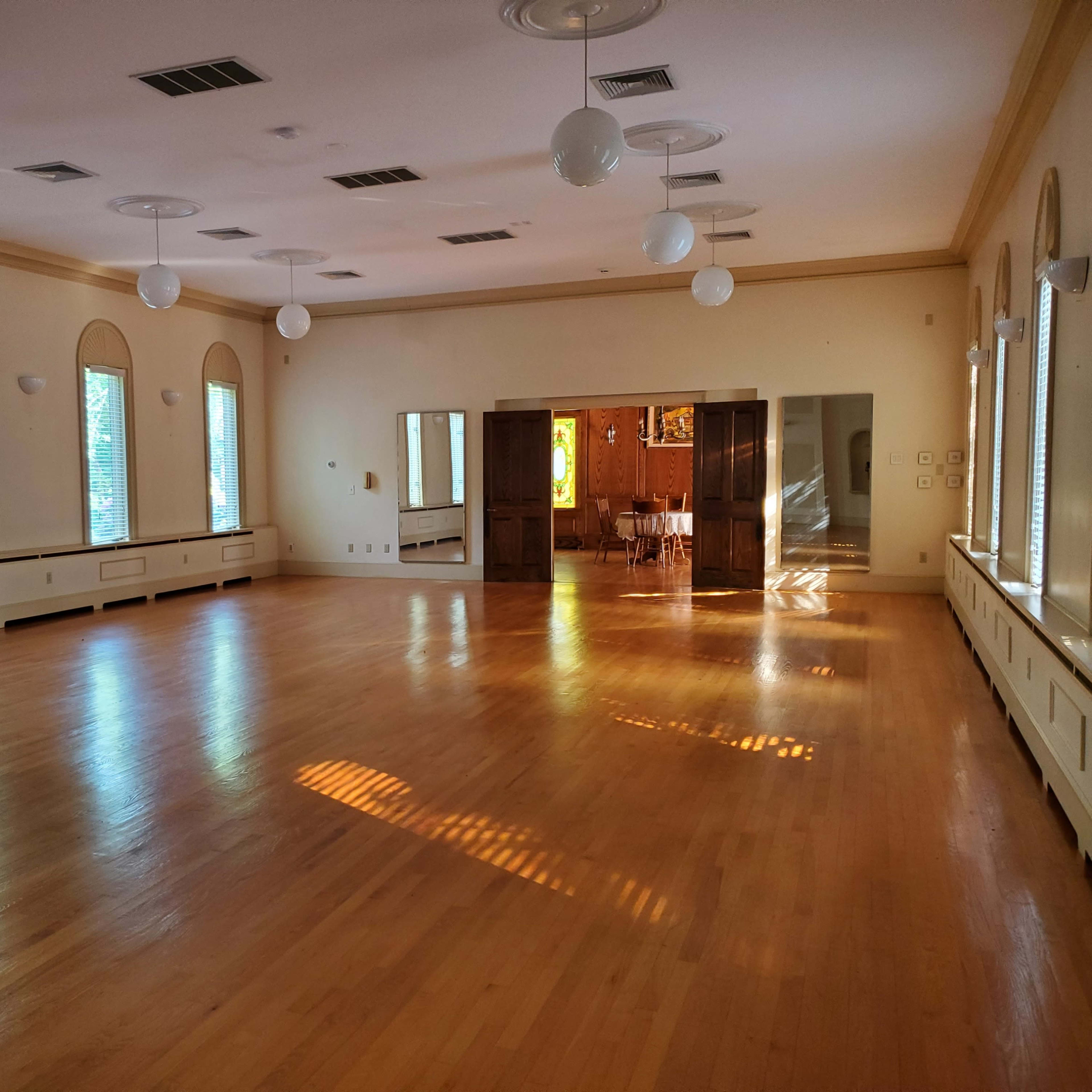 A spacious, well-lit room with hardwood floors, large windows, and a set of mirrors along one wall.