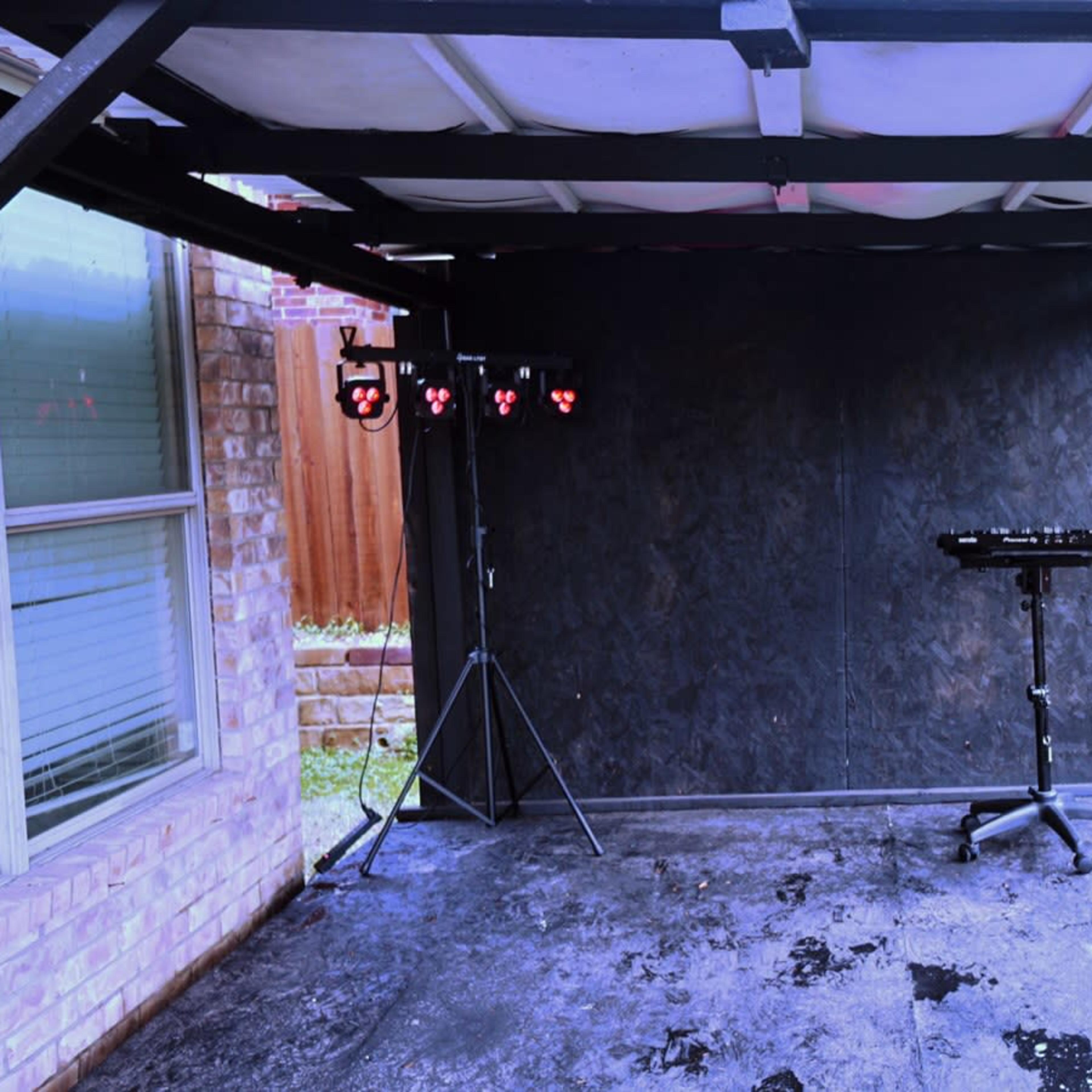 An empty outdoor stage set up with a black backdrop, a lighting rig on a stand, and a keyboard stand, surrounded by a grassy area and a wooden fence.