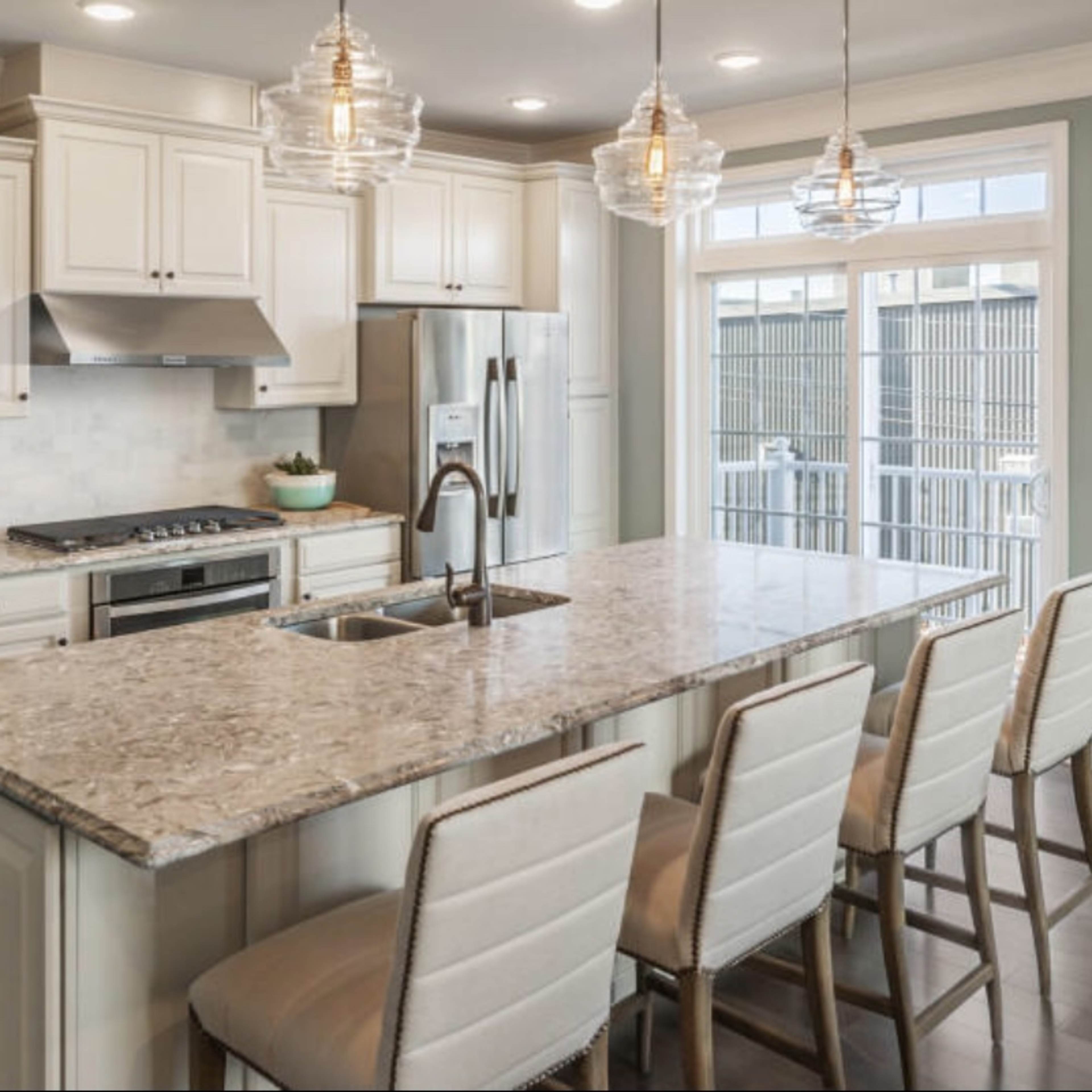 A modern kitchen with a large granite island, six beige bar stools, and stainless steel appliances.
