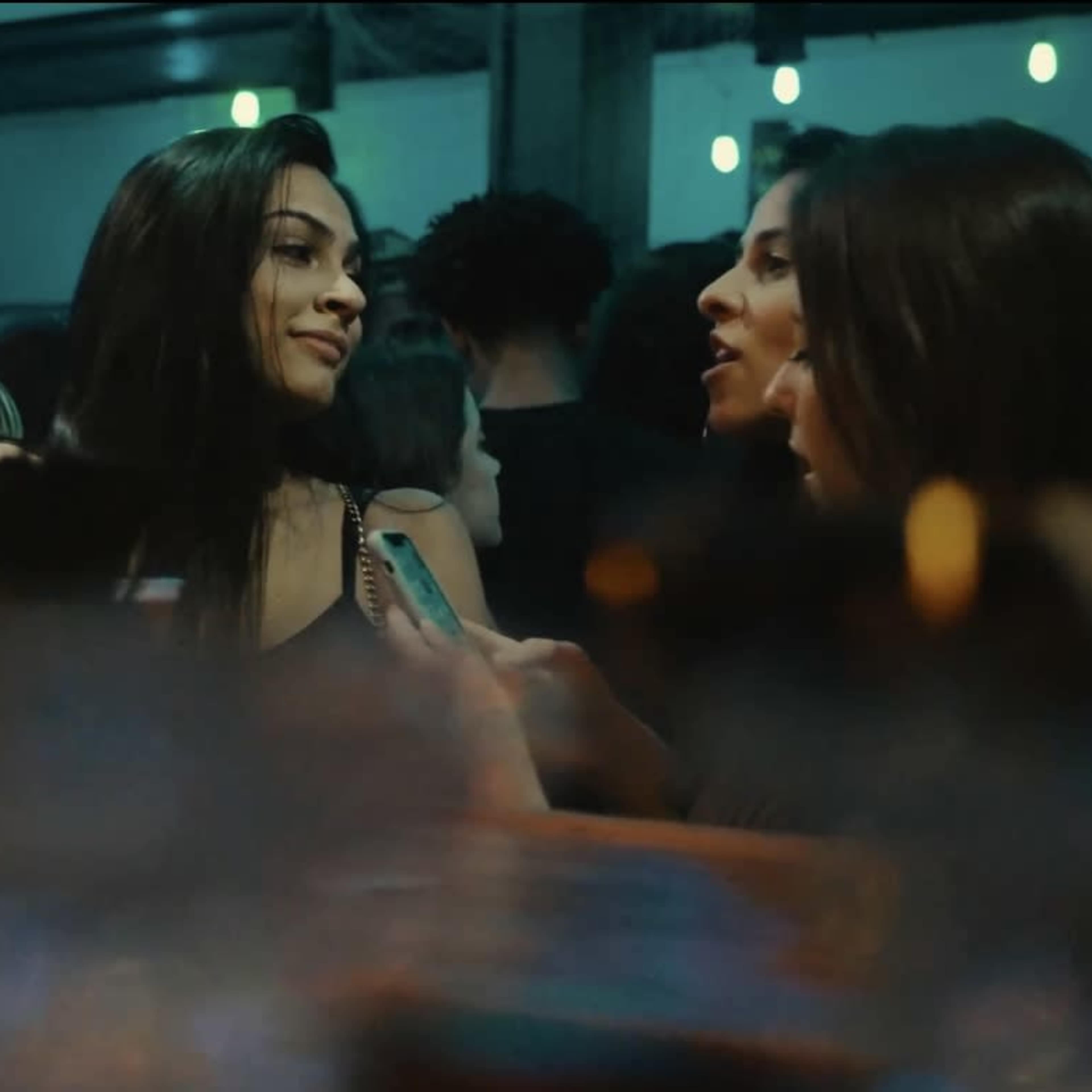 A lively bar scene features a crowd of people with two women engaging in conversation near the foreground.