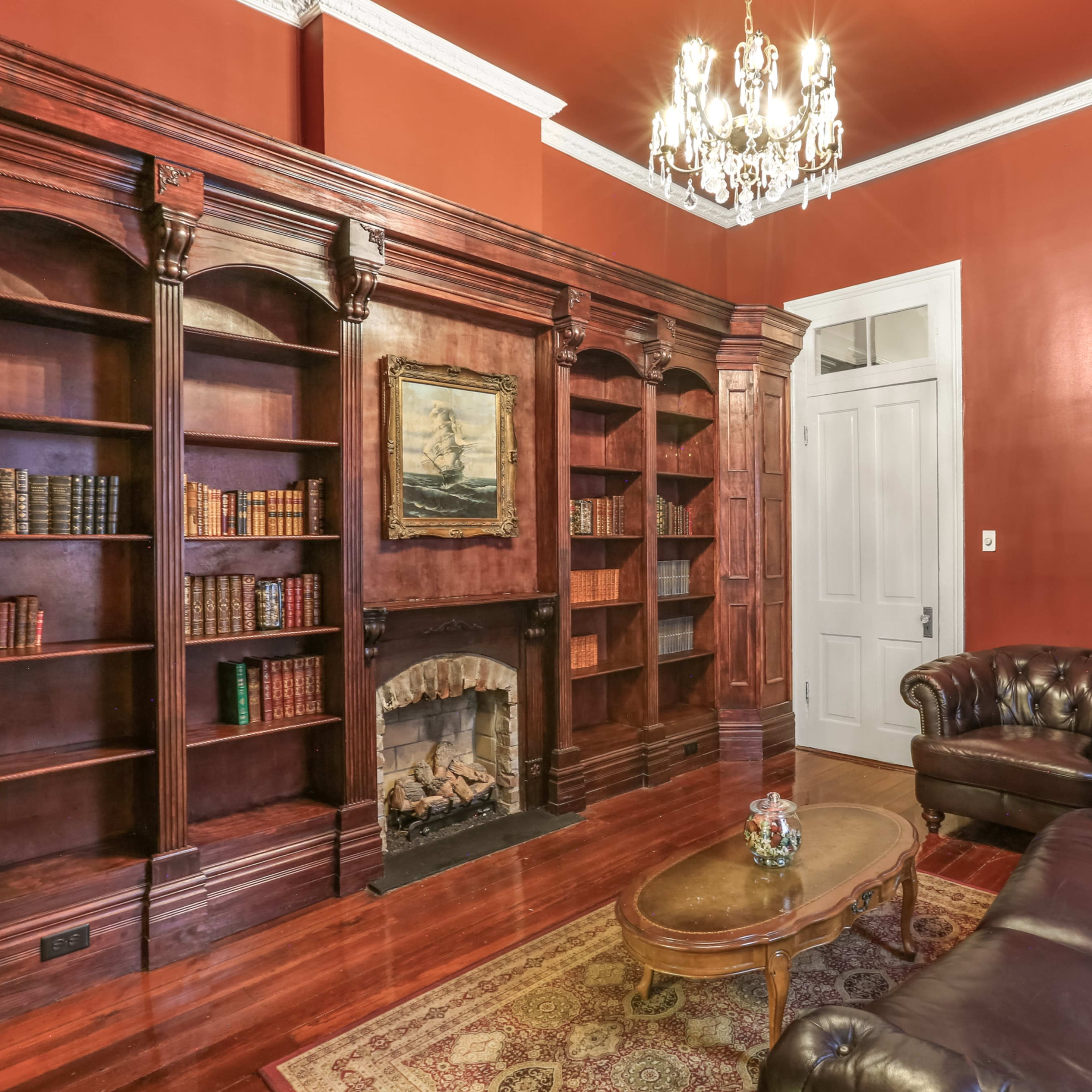 A richly furnished room with dark wood bookshelves, a leather chesterfield sofa, and a decorative chandelier against a red wall.