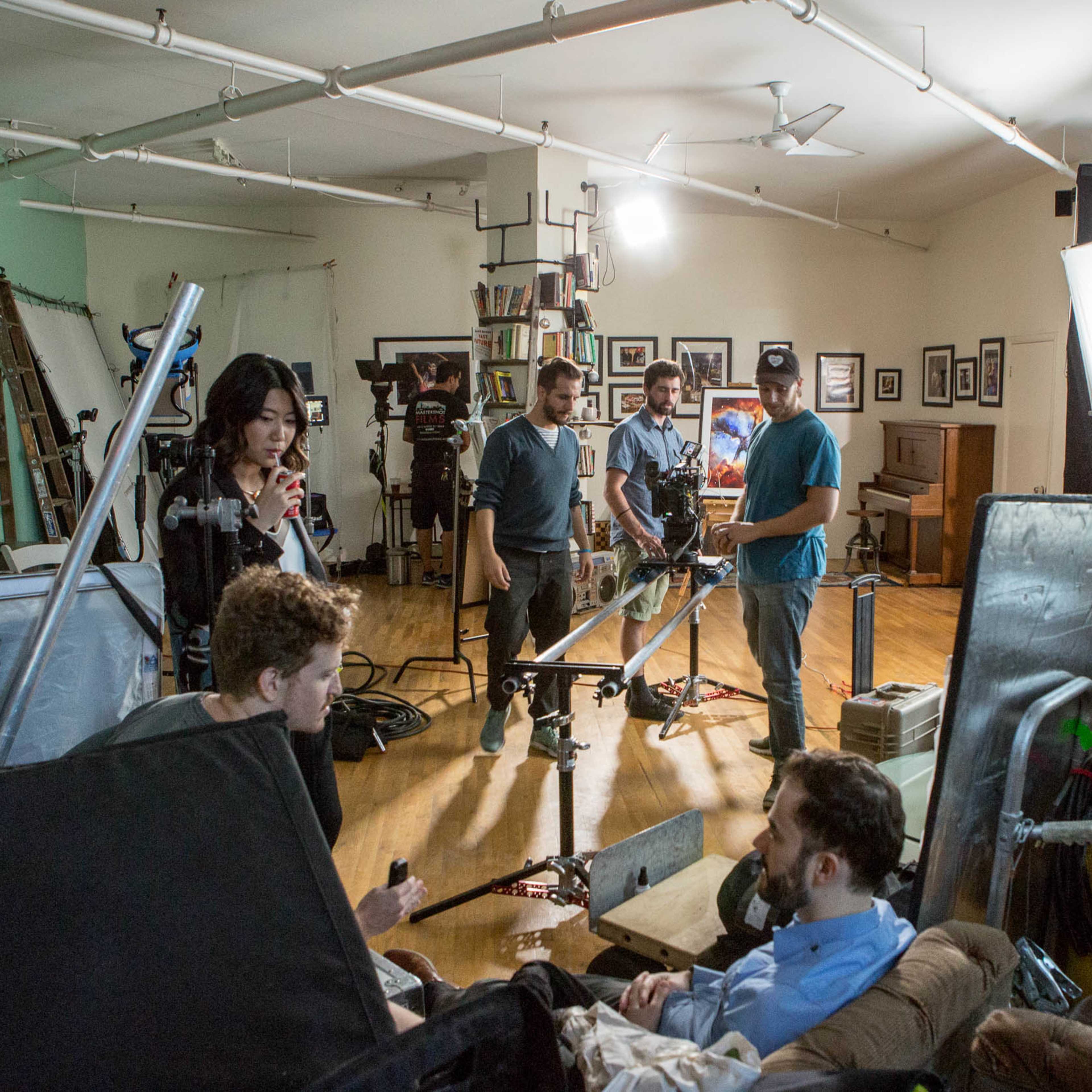 A film crew is set up in a studio, with actors and equipment arranged around a brightly lit scene.