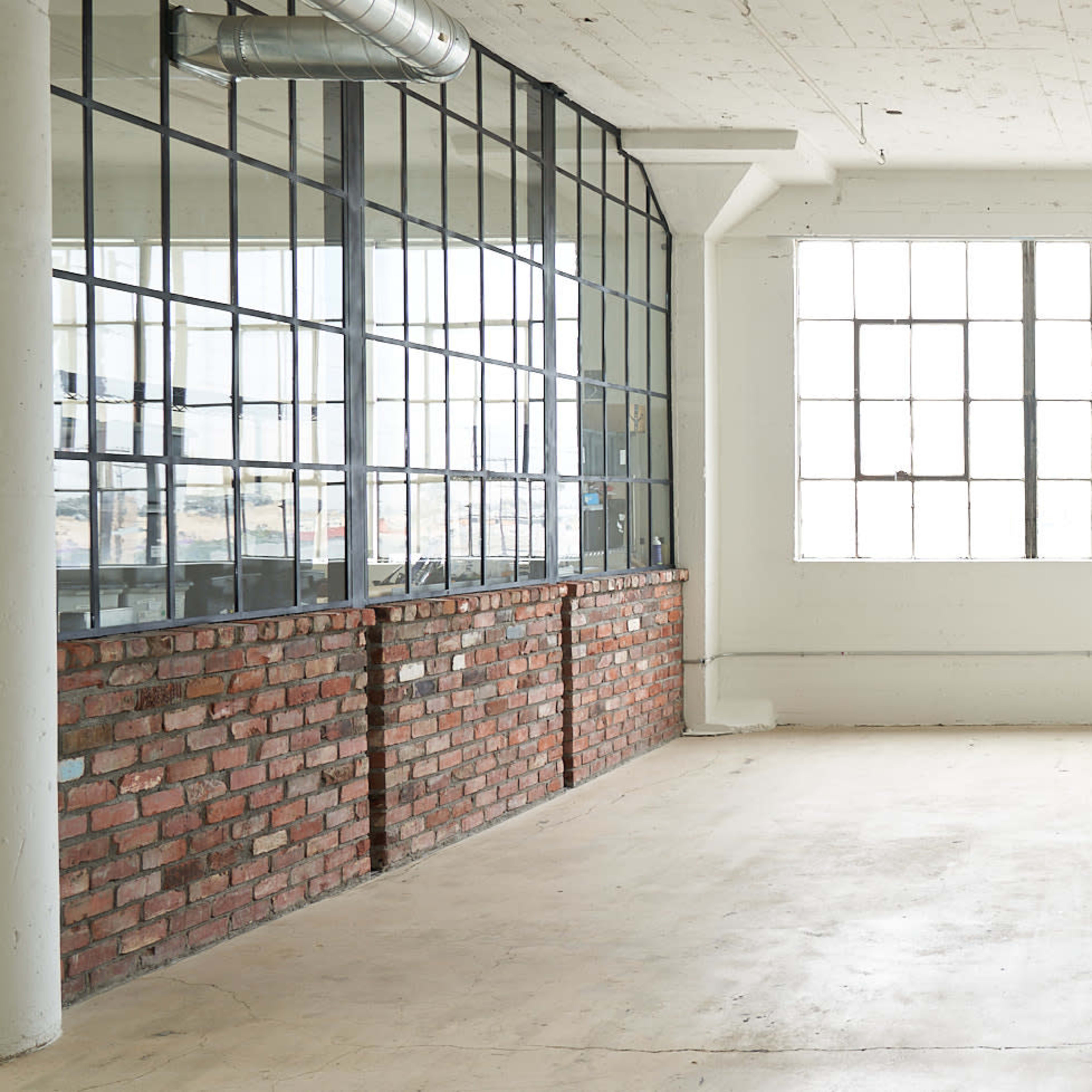 An empty interior space with large windows, exposed brick walls, and minimalistic furnishings.