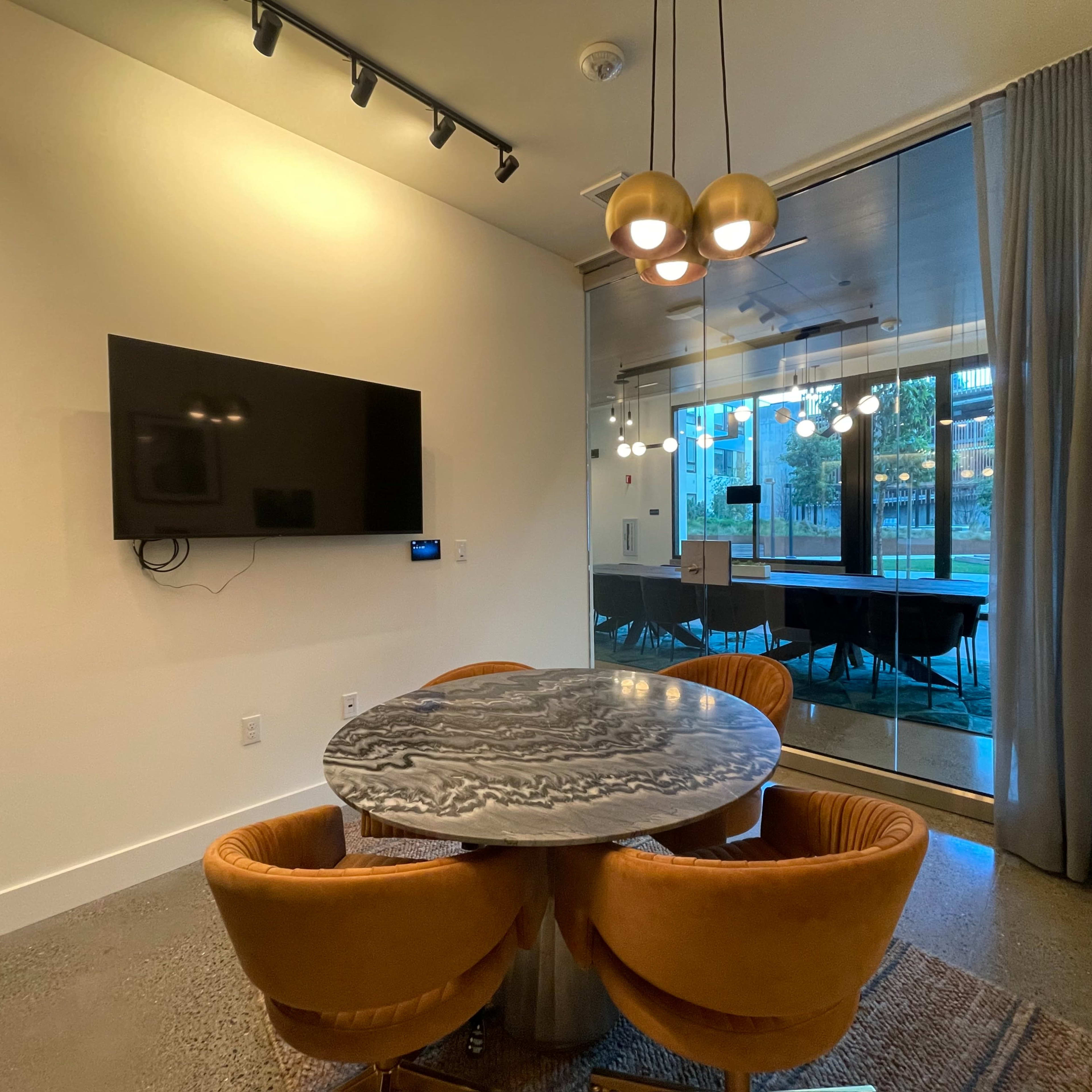 A modern meeting room features a round table with orange chairs, a large television mounted on the wall, and glass doors leading to an outdoor area.