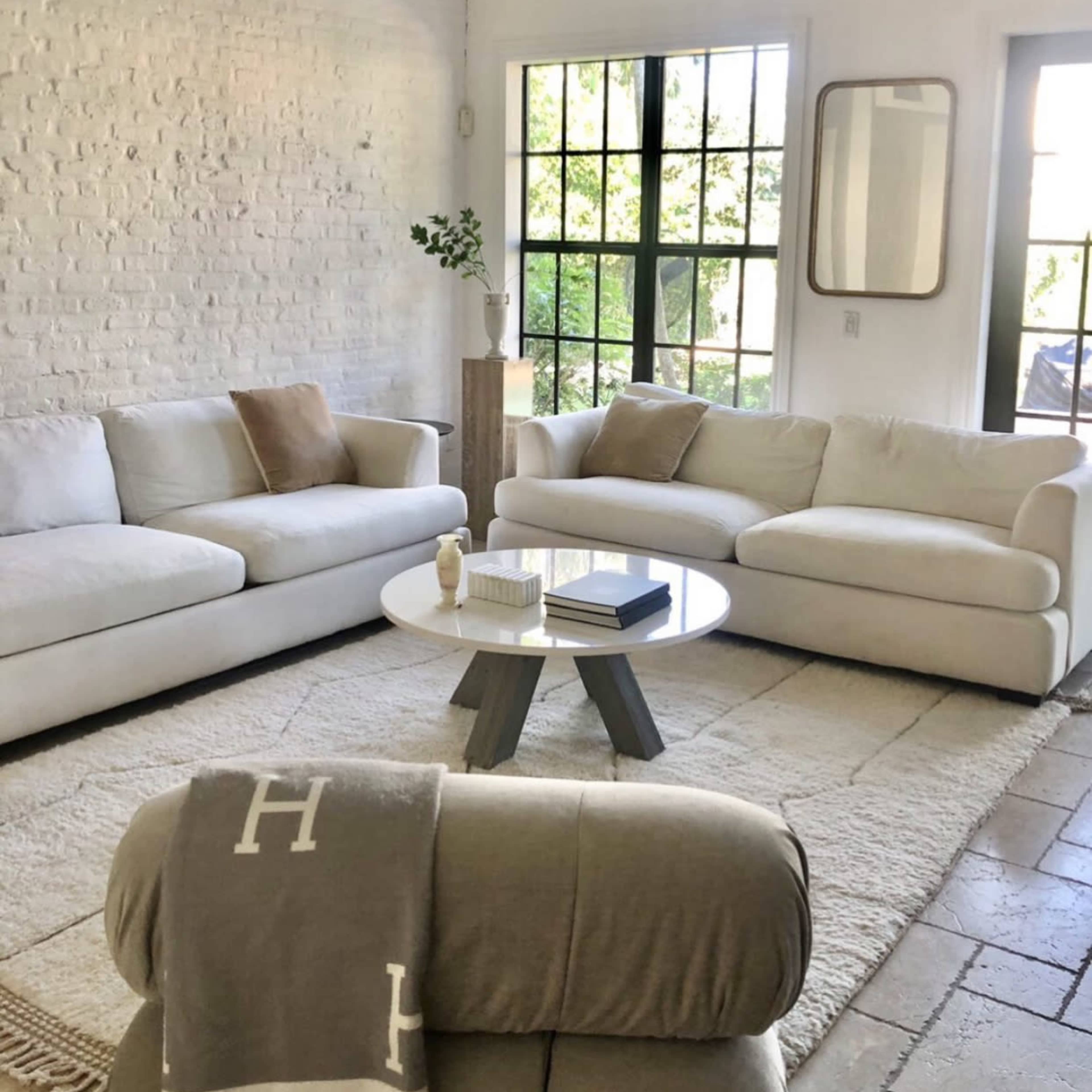 A bright, modern living room features two white sofas, a round coffee table, and large windows that allow natural light to fill the space.