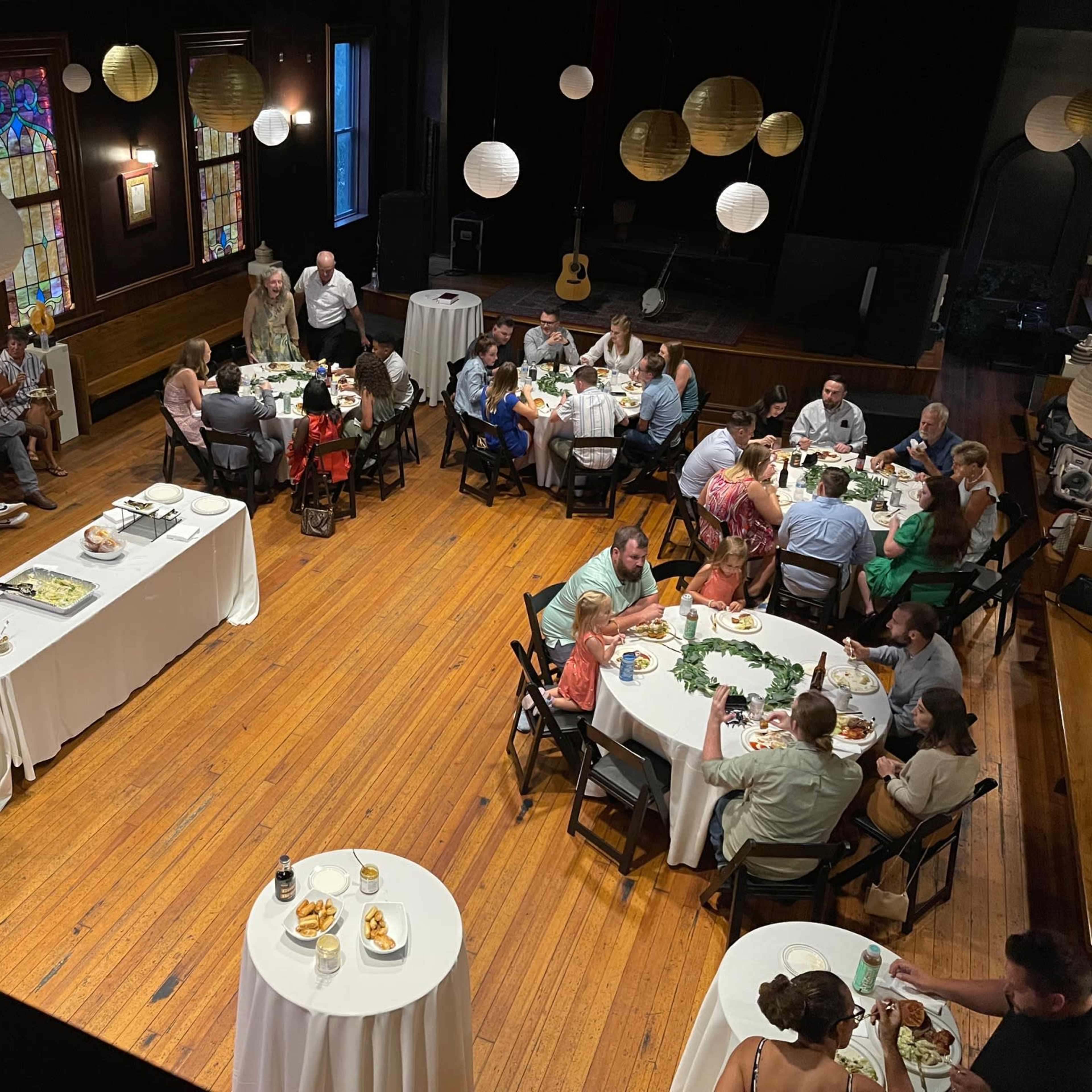 A gathering is taking place in a dining hall with several tables set up for guests enjoying a meal.
